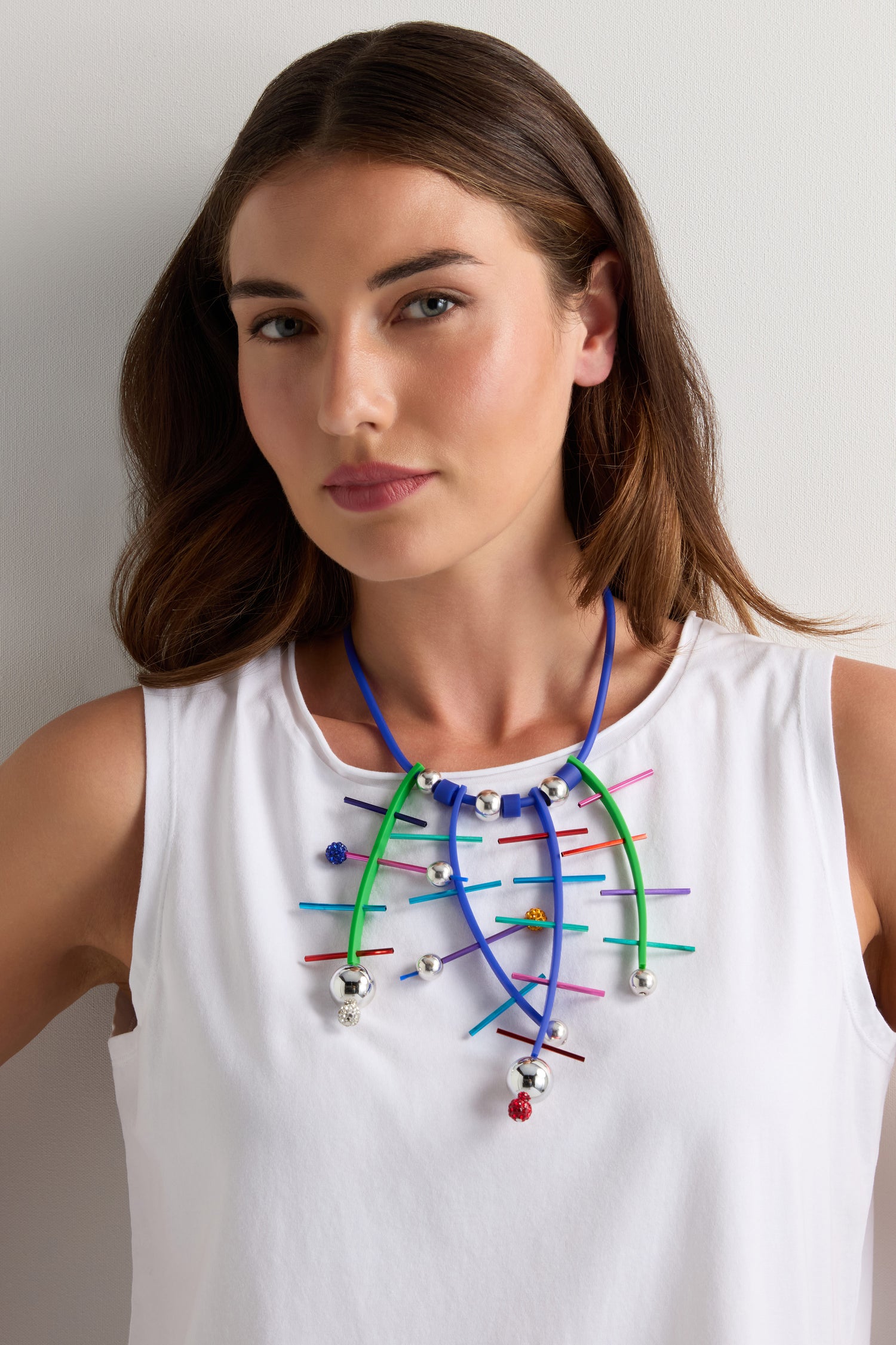 A woman in a white sleeveless top models the Fame Necklace, a handmade Parisian piece crafted from vibrant blue, green, and pink flexible tubes with silver bells and playful pom-poms.