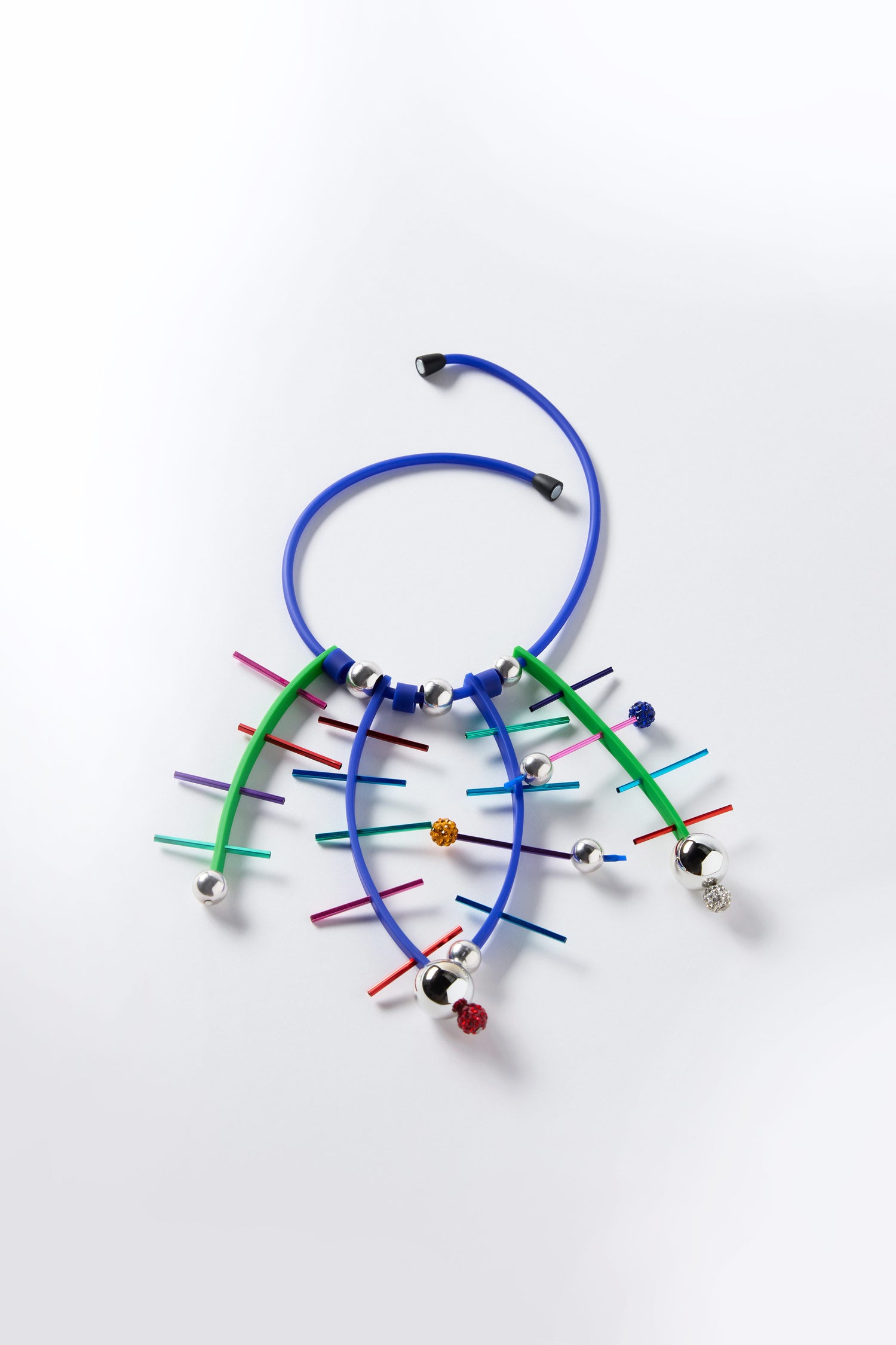 The Fame Necklace by Samuel Coraux is handmade in Paris with blue and green flexible tubes, silver spheres, and multicolored rods for a vibrant spiky look, shown on a white background.
