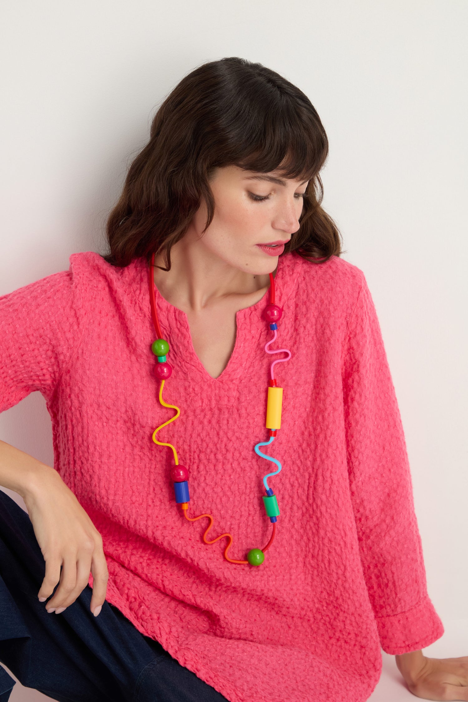 A woman in a textured pink top looks down, wearing the Rainbow Graphic Pop Necklace—sculptural jewelry featuring chunky, colorful beads—set against a plain light background.