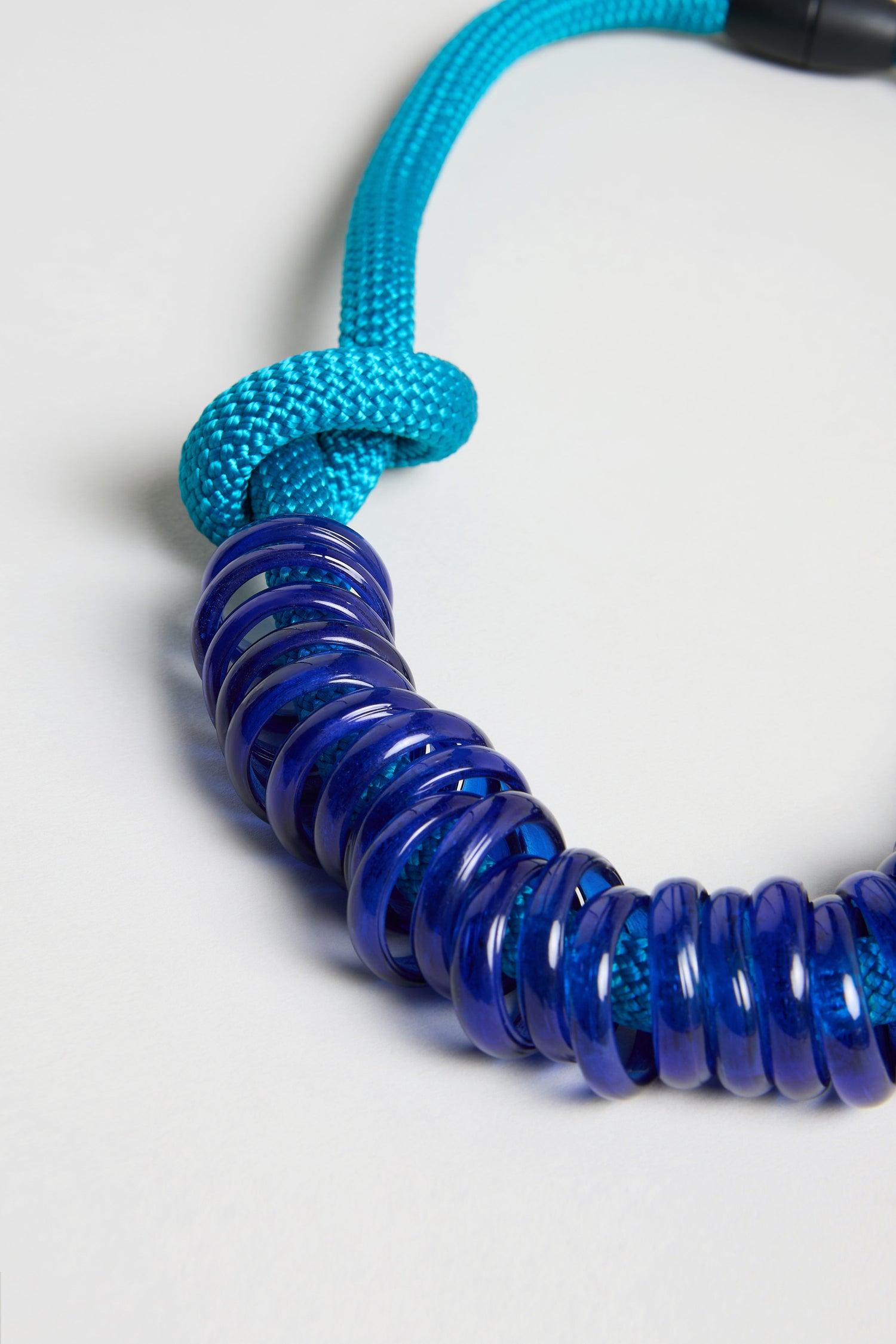 A close-up of the Murano Glass Rings Necklace: a blue braided cord necklace with several translucent dark blue glass rings.