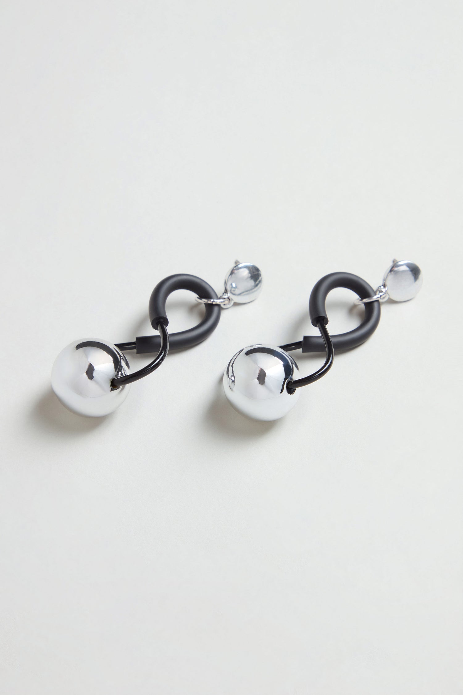 The Infinity Earrings display a sculptural design with black curved links connected to polished silver spheres and smaller silver bead studs, all set against a light background.