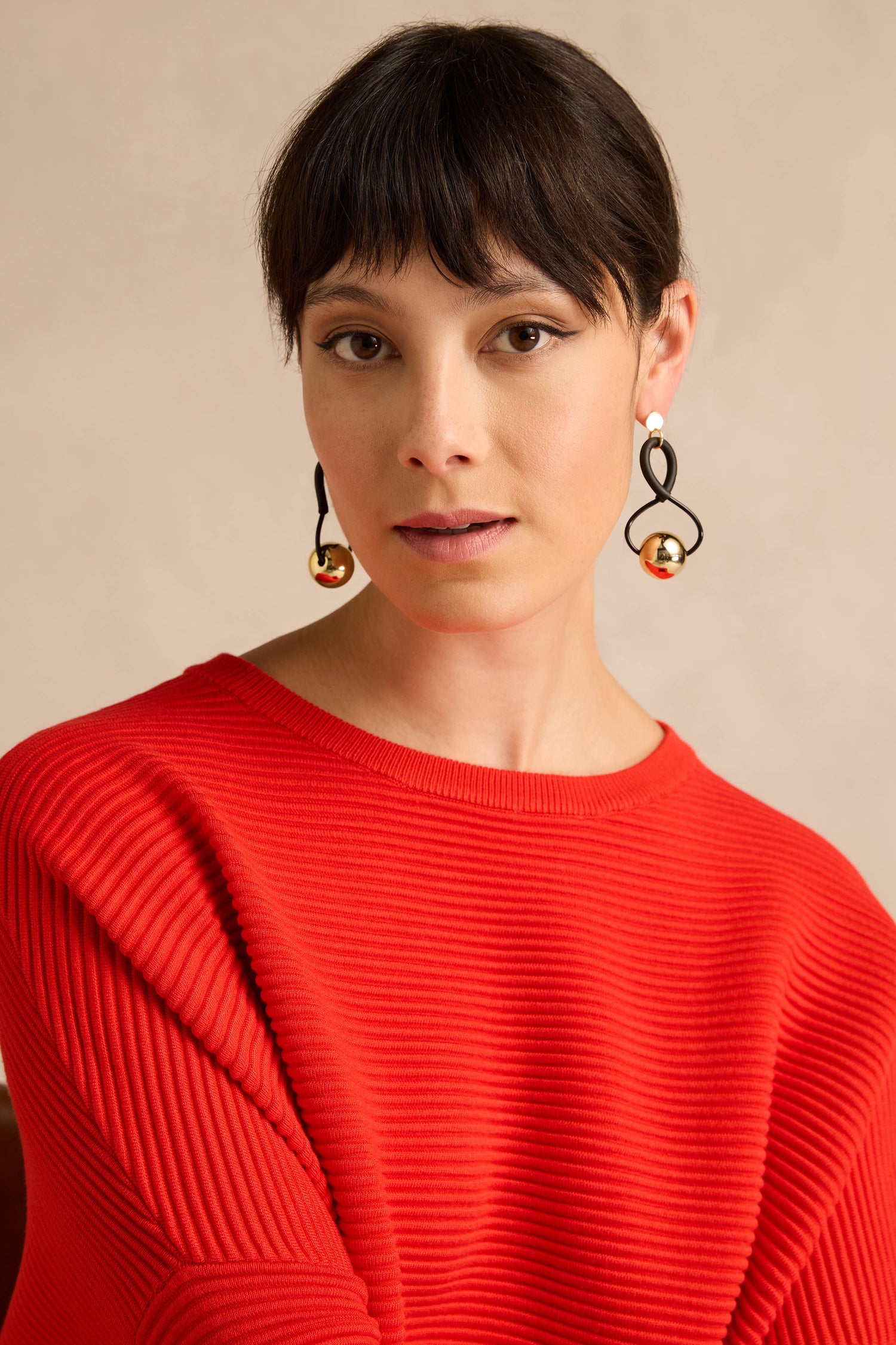 Woman with short dark hair wearing a bright red ribbed sweater and striking Infinity Earrings—handmade in France, featuring a black-and-gold sculptural design—posing against a neutral background.