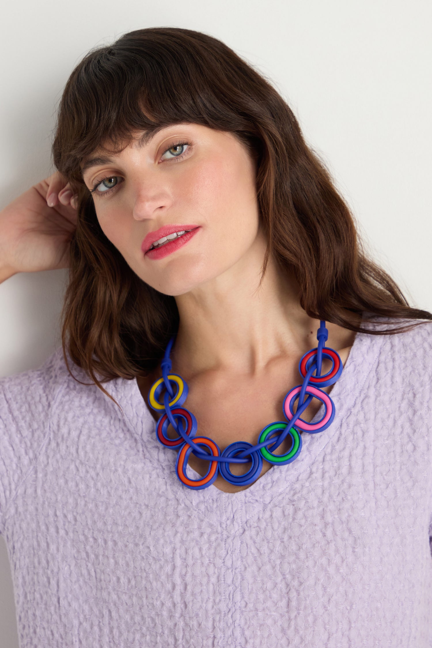 A woman with medium-length brown hair models the Colour Pop Rings Necklace, a handmade Parisian piece with interlocking rings, while wearing a textured light purple top and posing with one hand behind her head against a plain background.