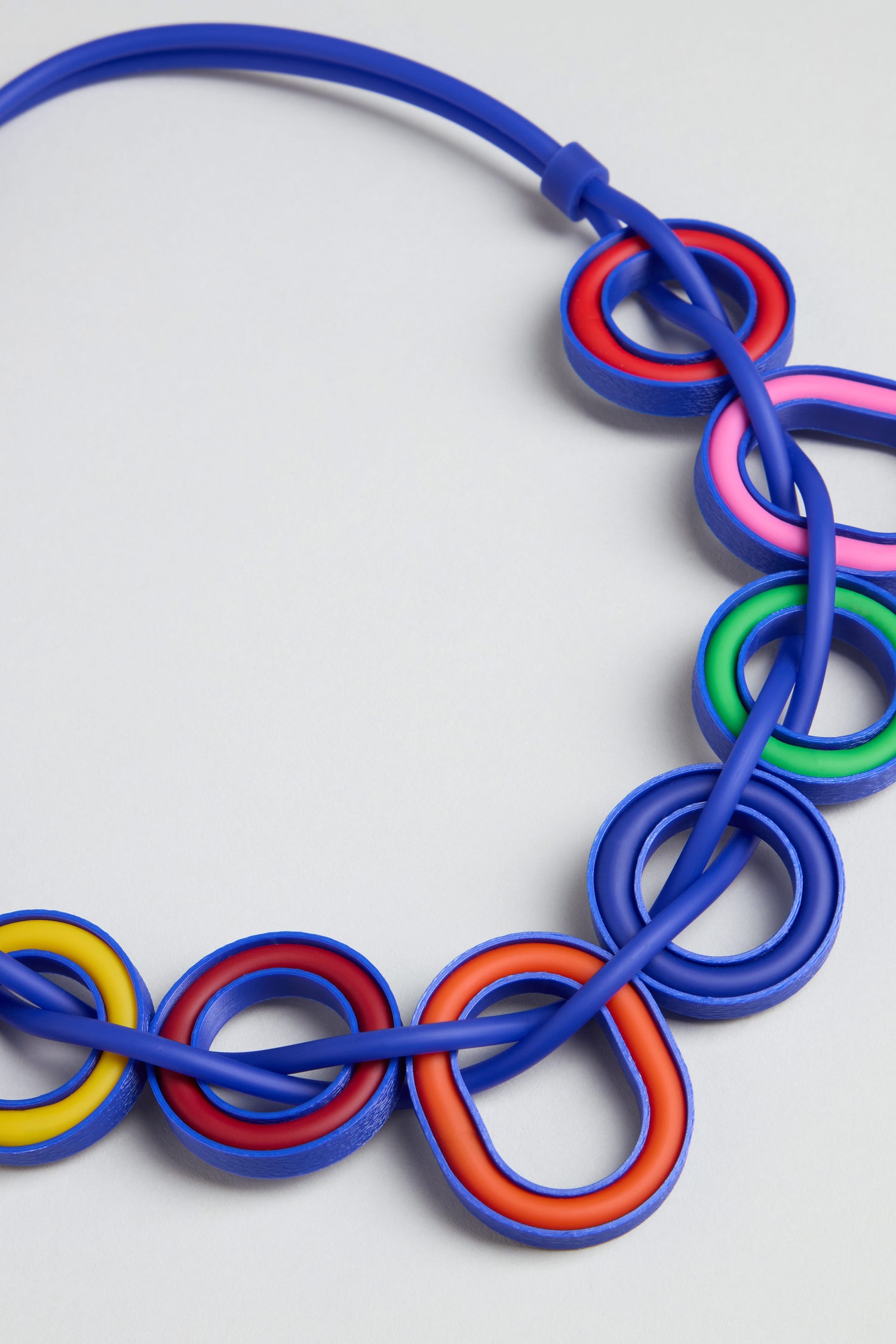 The Colour Pop Rings Necklace features interlocking blue rings accented by vibrant red, yellow, pink, and green ovals, all beautifully set against a light gray background.
