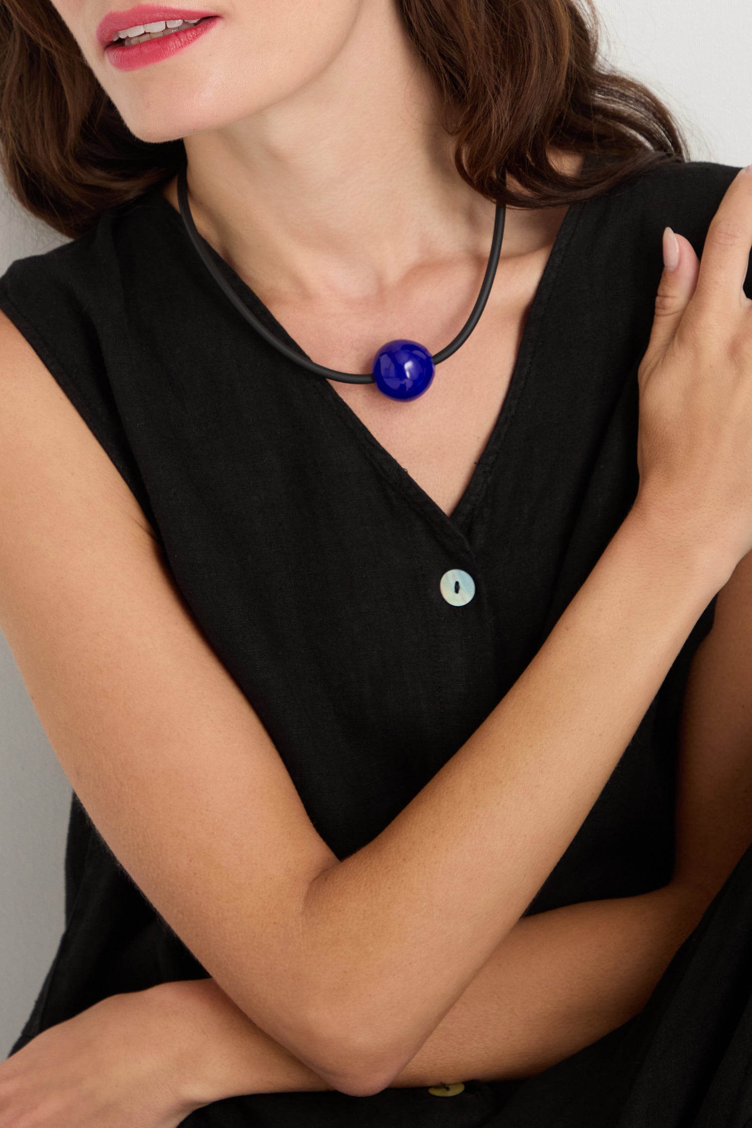 A woman in a sleeveless black V-neck dress wears the Murano Bead Necklace, handmade in Paris, shown from shoulders to mouth with arms crossed.
