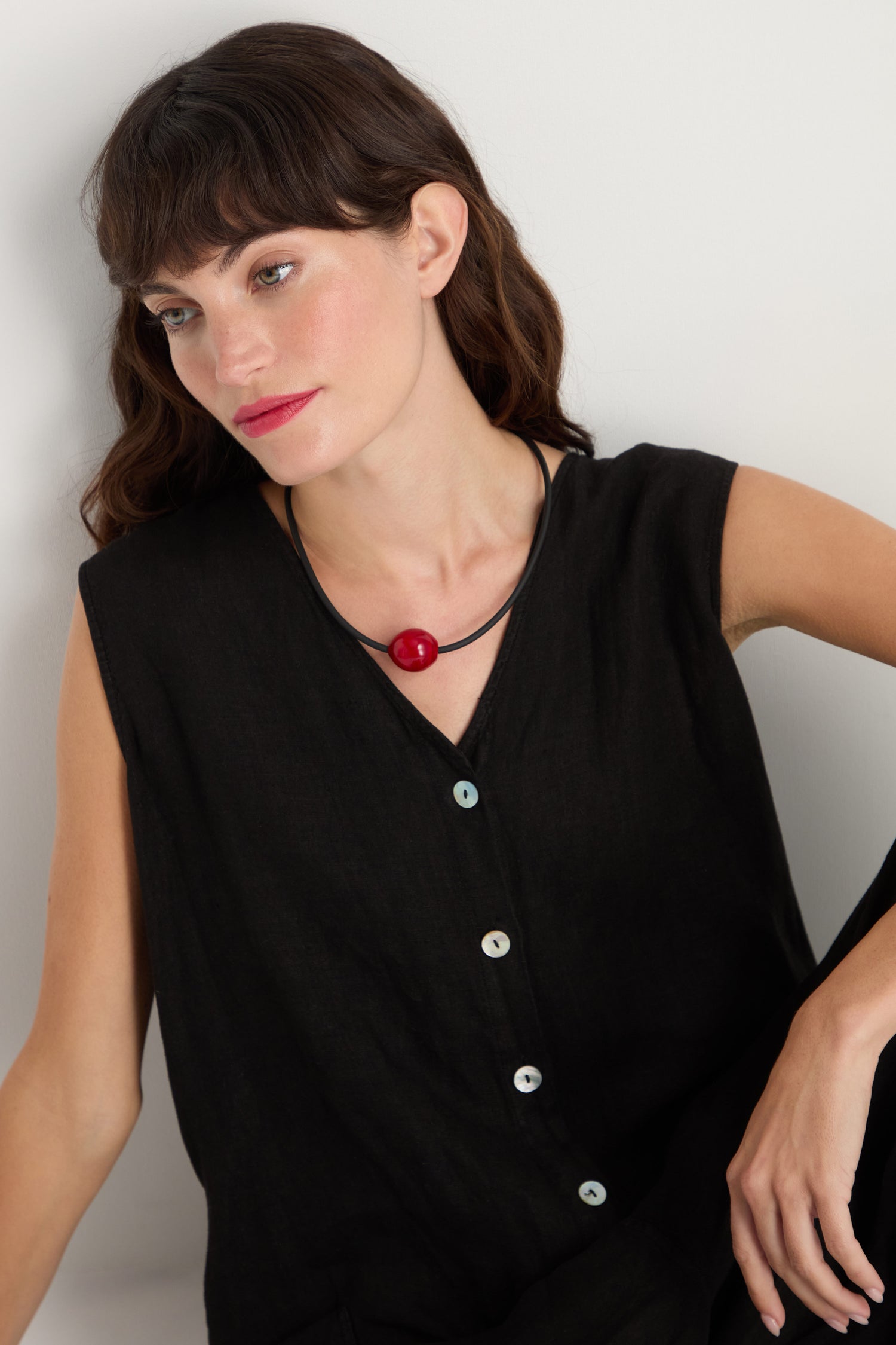 Woman with wavy brown hair wears a sleeveless black top and the Murano Bead Necklace, featuring a large red bead, handmade in Paris, while sitting against a white background.