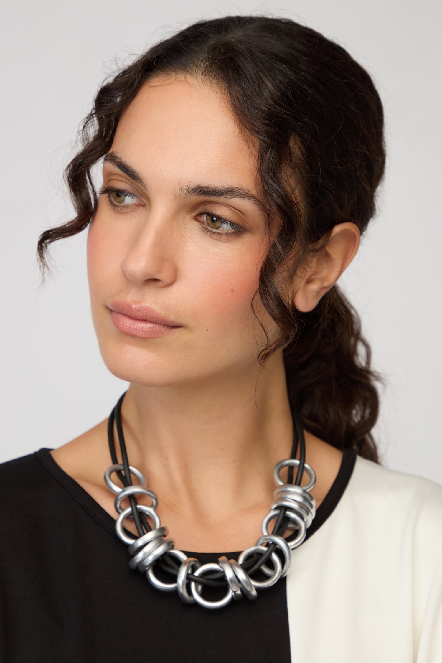 A woman with wavy brown hair in a black and white top models the Black and Silver Metal Rings Necklace by Samuel Coraux, a contemporary statement piece, as she looks slightly to the side against a plain background.