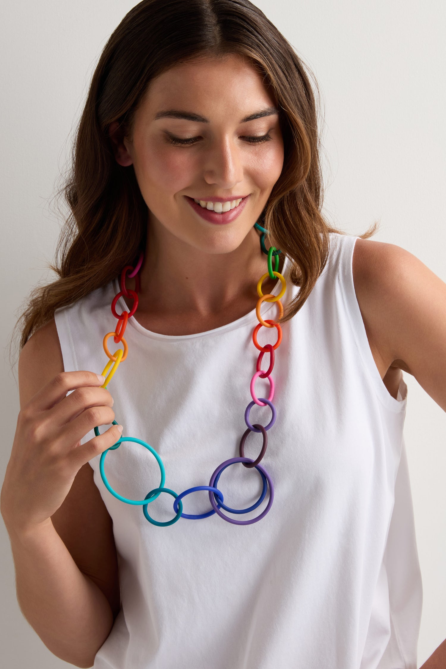 Woman wearing a white sleeveless top and the Long Rainbow Hoops Necklace by Samuel Coraux, looking down and smiling against a plain background.