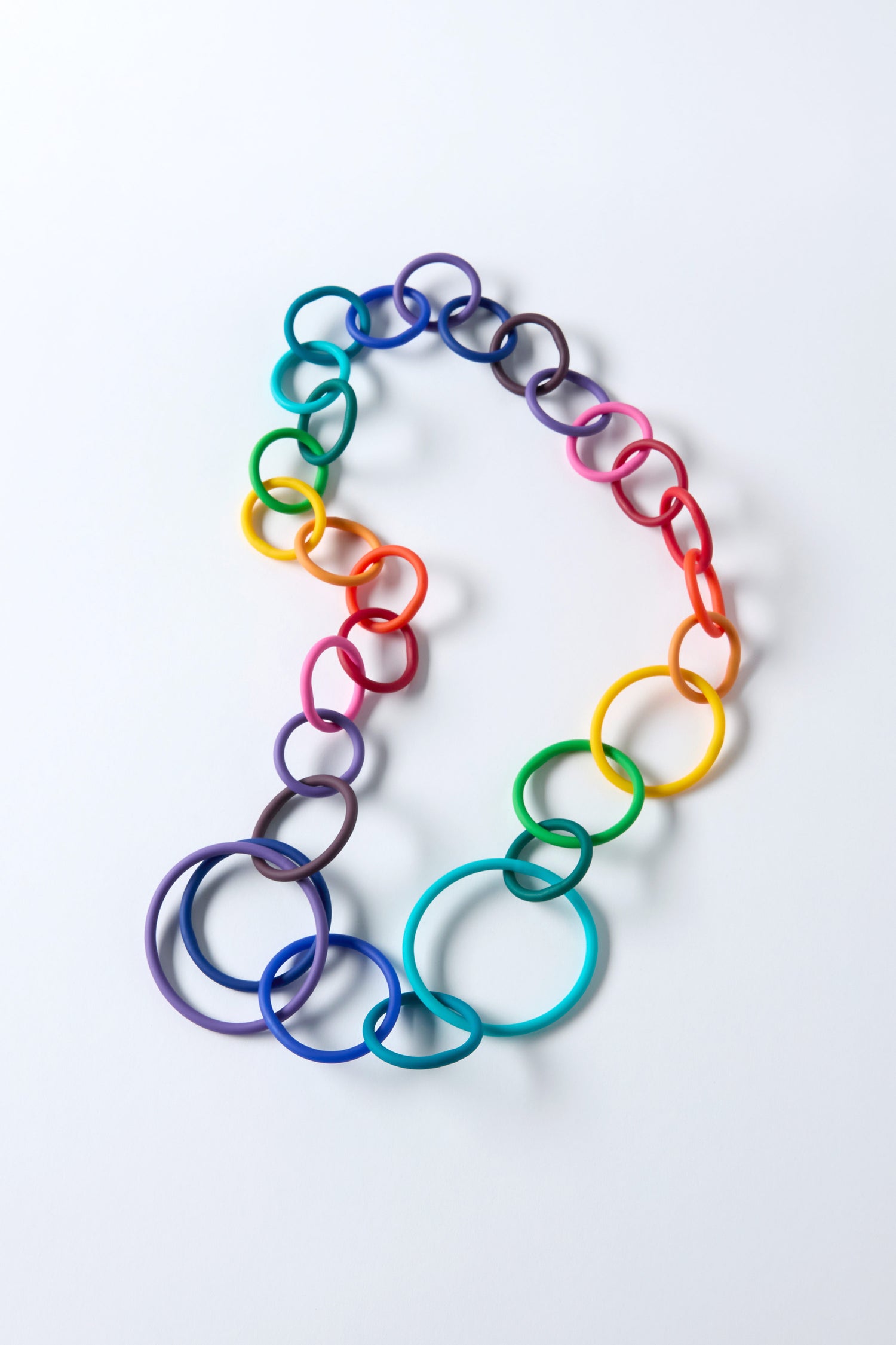 The Long Rainbow Hoops Necklace by Samuel Coraux showcases interlocking multicolored rubber hoops, artfully arranged in a gradient from purple to red on a white background.