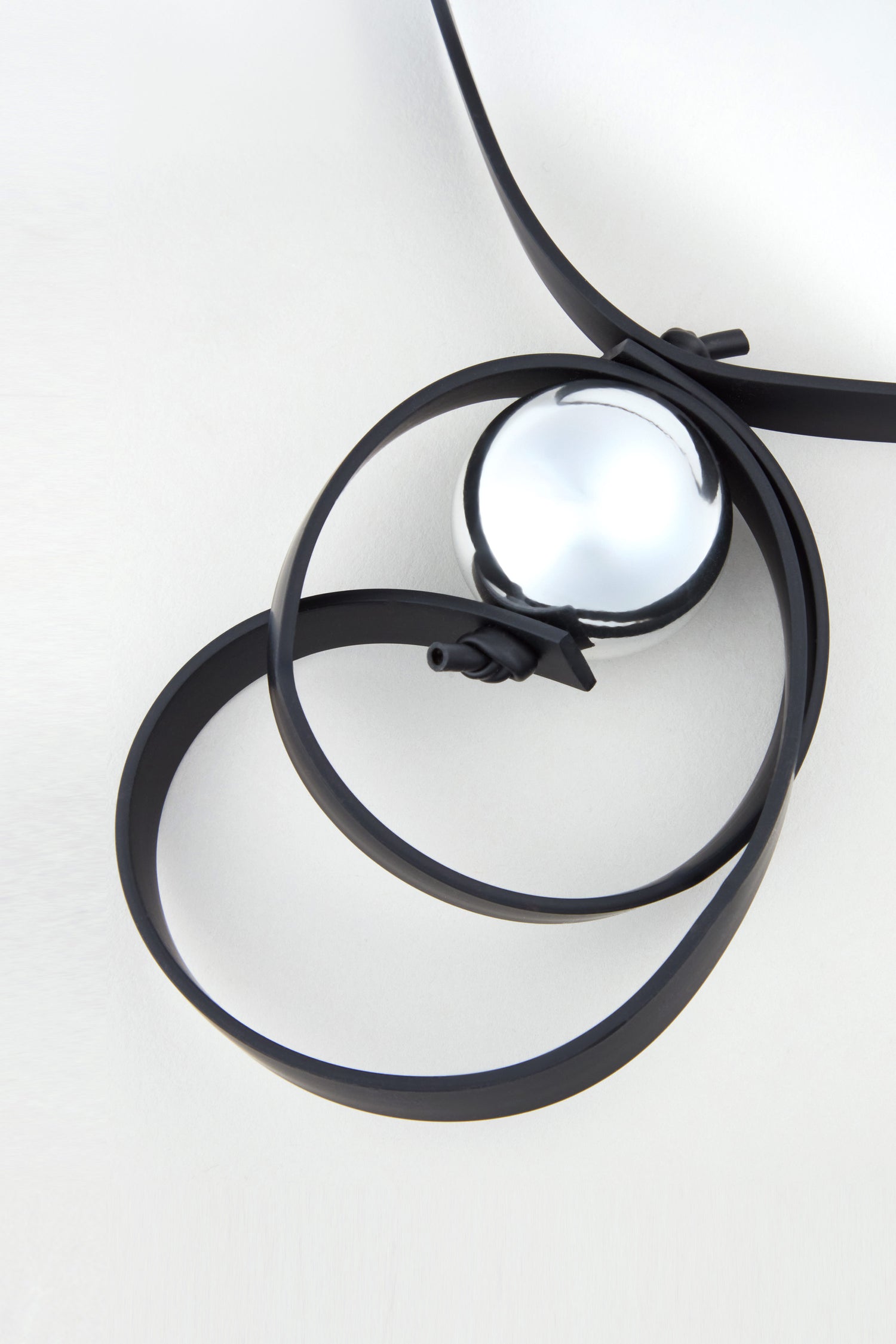 A black leather belt is loosely coiled around the shiny Penseur Silver Sphere Necklace on a plain white surface, highlighting the bold spirit of sculptural jewellery.