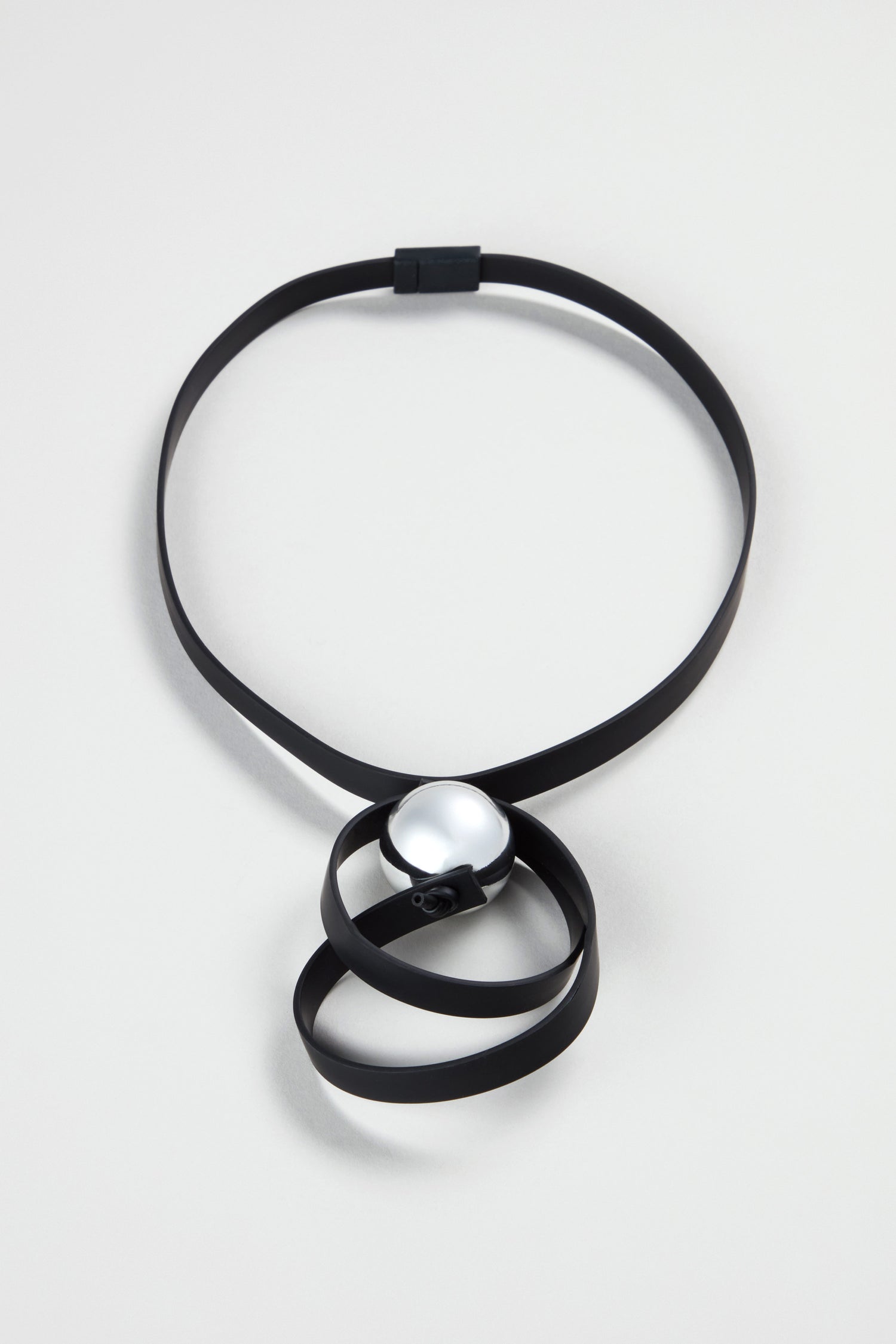 The Penseur Silver Sphere Necklace features a flat black band with a large metallic silver bead and abstract loops at the center, showcasing Samuel Coraux’s signature sculptural style, displayed against a light background.