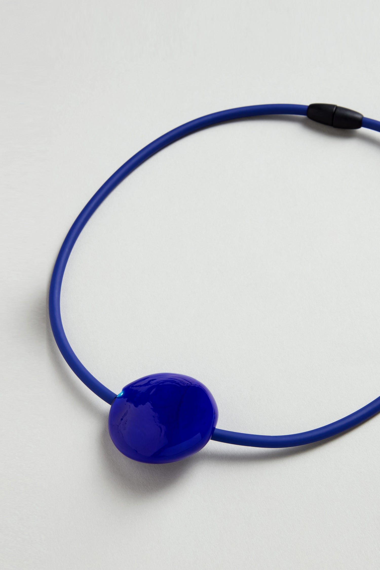The Murano Glass Pebble Necklace features a blue cord, glossy irregular blue pendant, and black clasp, all displayed on a light background.
