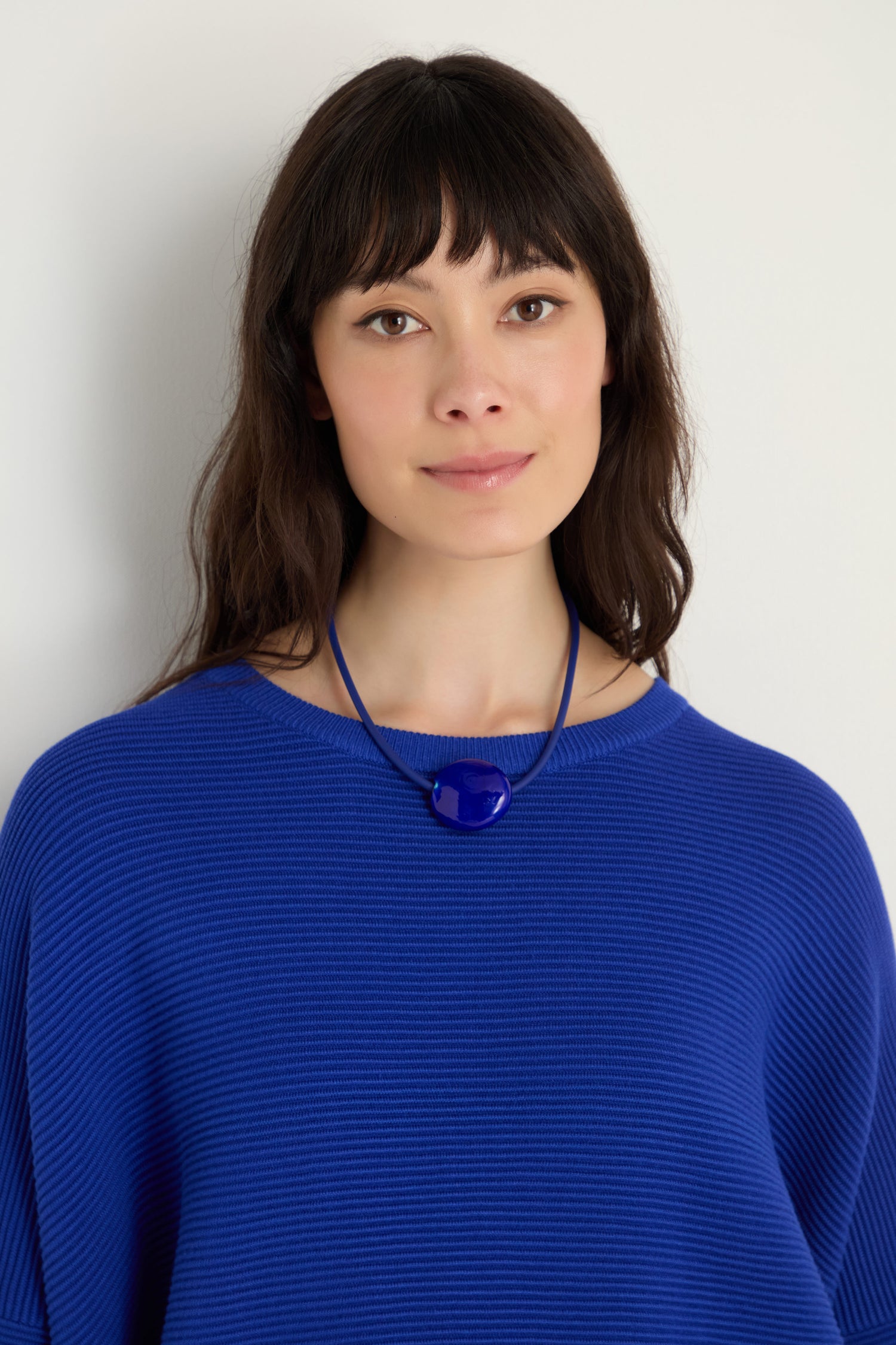 A woman with long dark hair, dressed in a blue top and wearing the Murano Glass Pebble Necklace, stands against a plain light background.