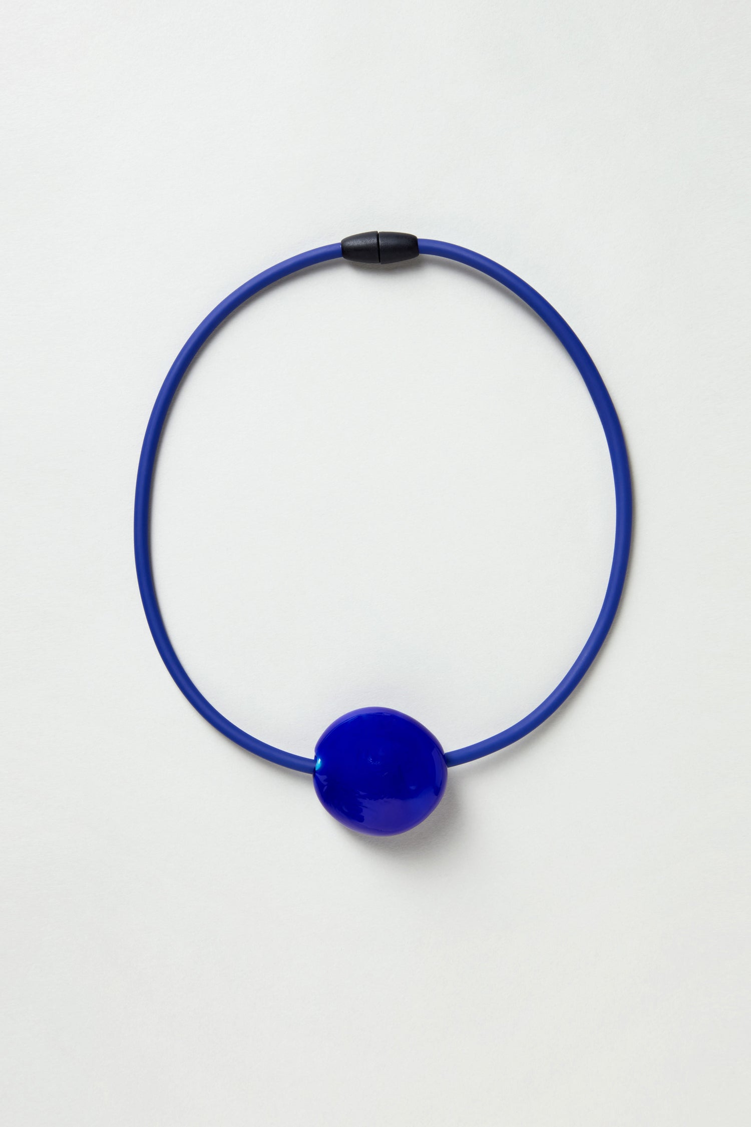 The Murano Glass Pebble Necklace features a blue circular design with a large, glossy oval bead at the center and a black clasp, shown on a white background.