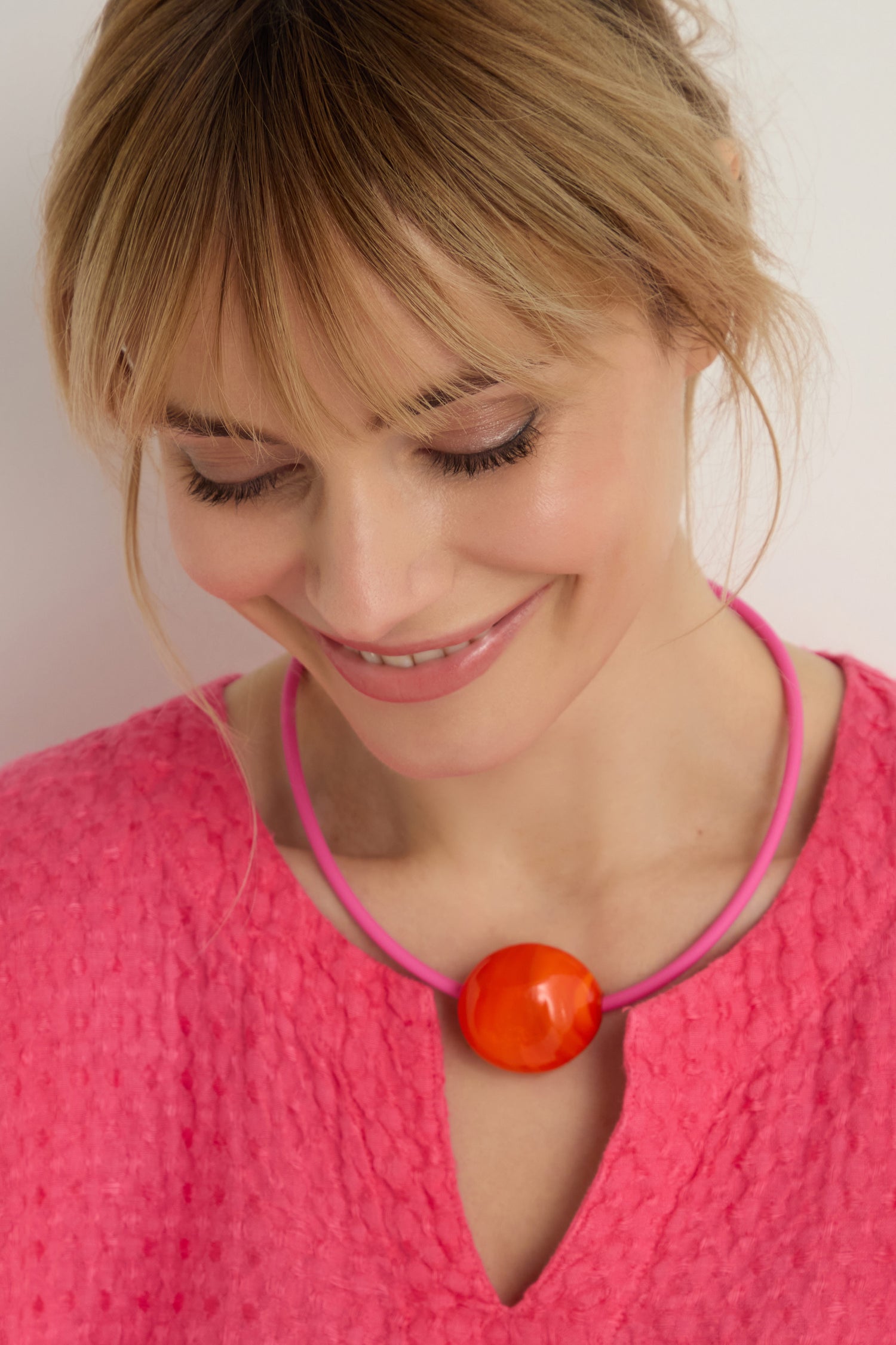 Woman with light brown hair wearing a textured pink top and the Murano Glass Pebble Necklace featuring a bold orange accent, looking down and smiling.