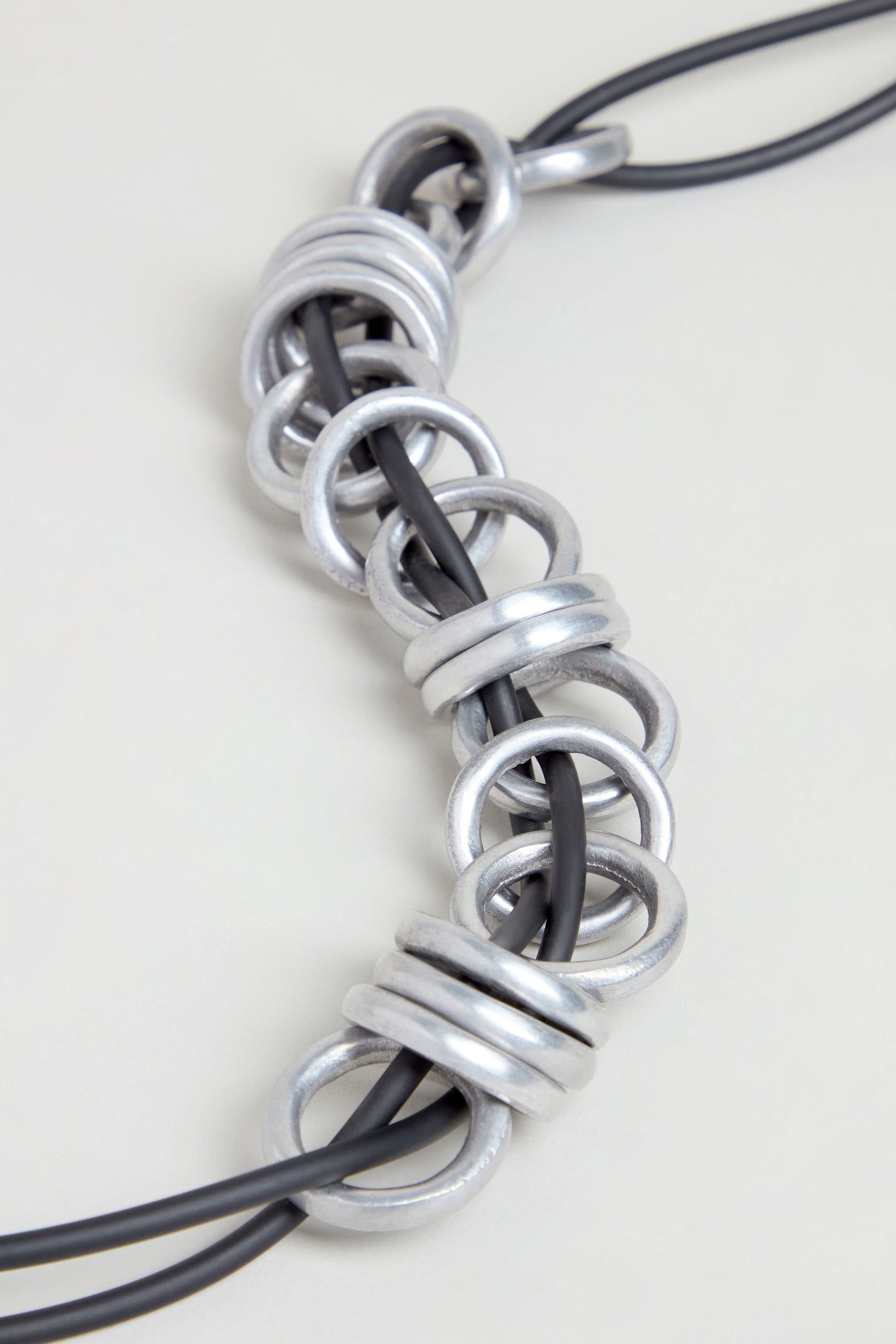 The Metal Rings Necklace features interlocking silver rings threaded onto black cords and arranged in a linear pattern on a light background, showcasing a sculptural design that is handmade in France.
