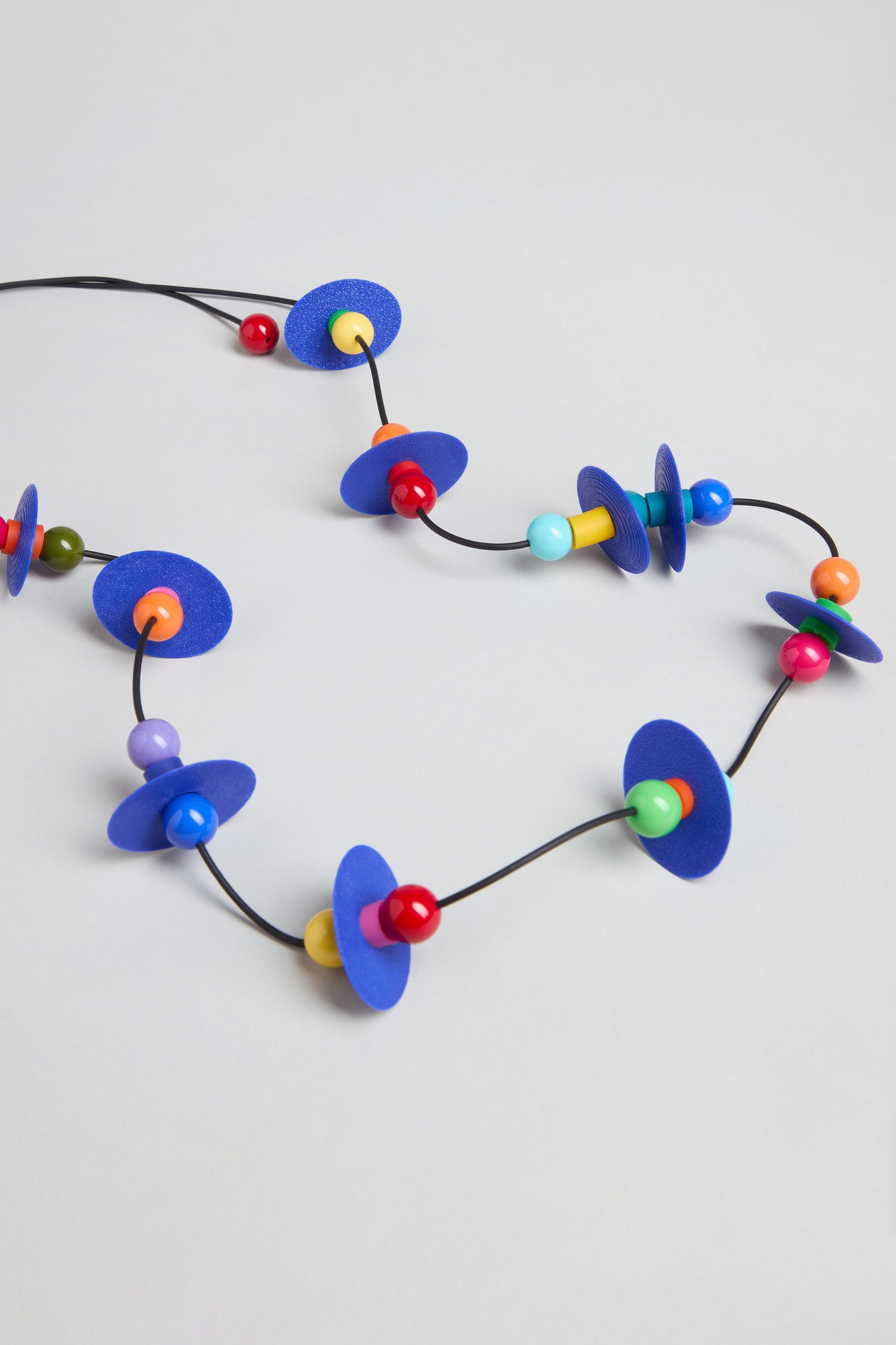 The Orbit Necklace showcases abstract blue flat discs and multicolored round beads on a black cord, arranged against a light gray background.