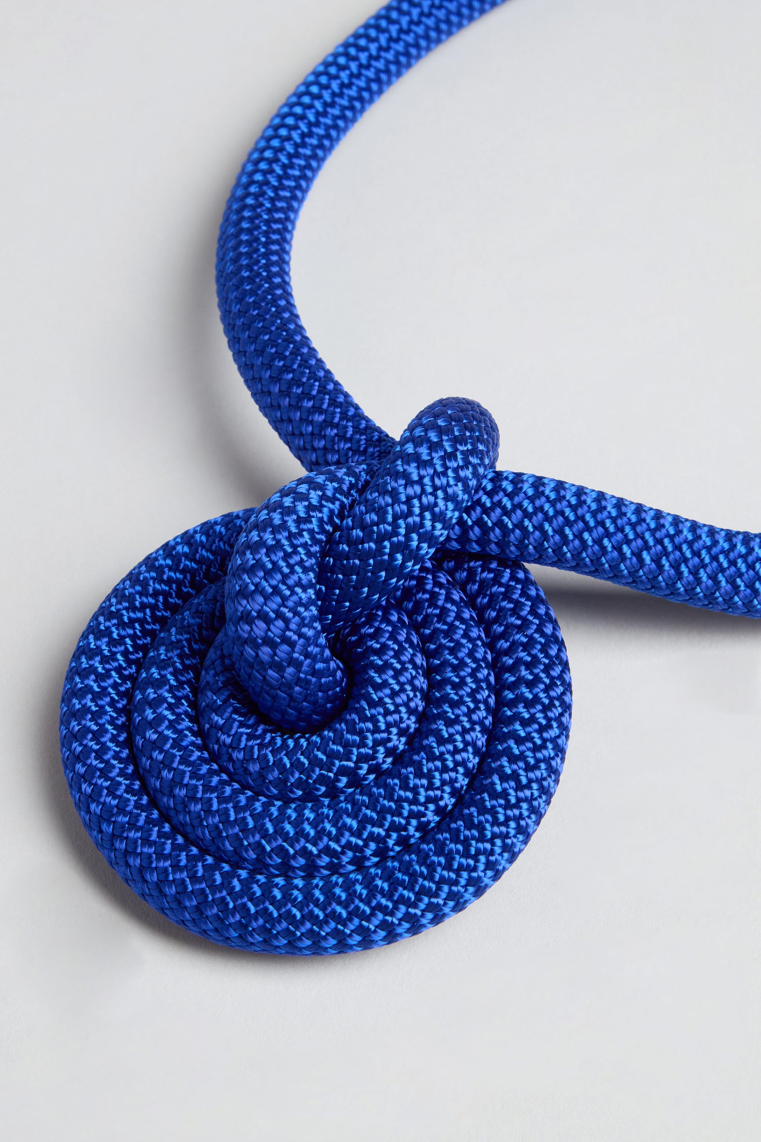 A close-up of the Spiral Cord Necklace, featuring a blue rope tied in a circular knot on a light gray surface, highlights its unique spiral design.
