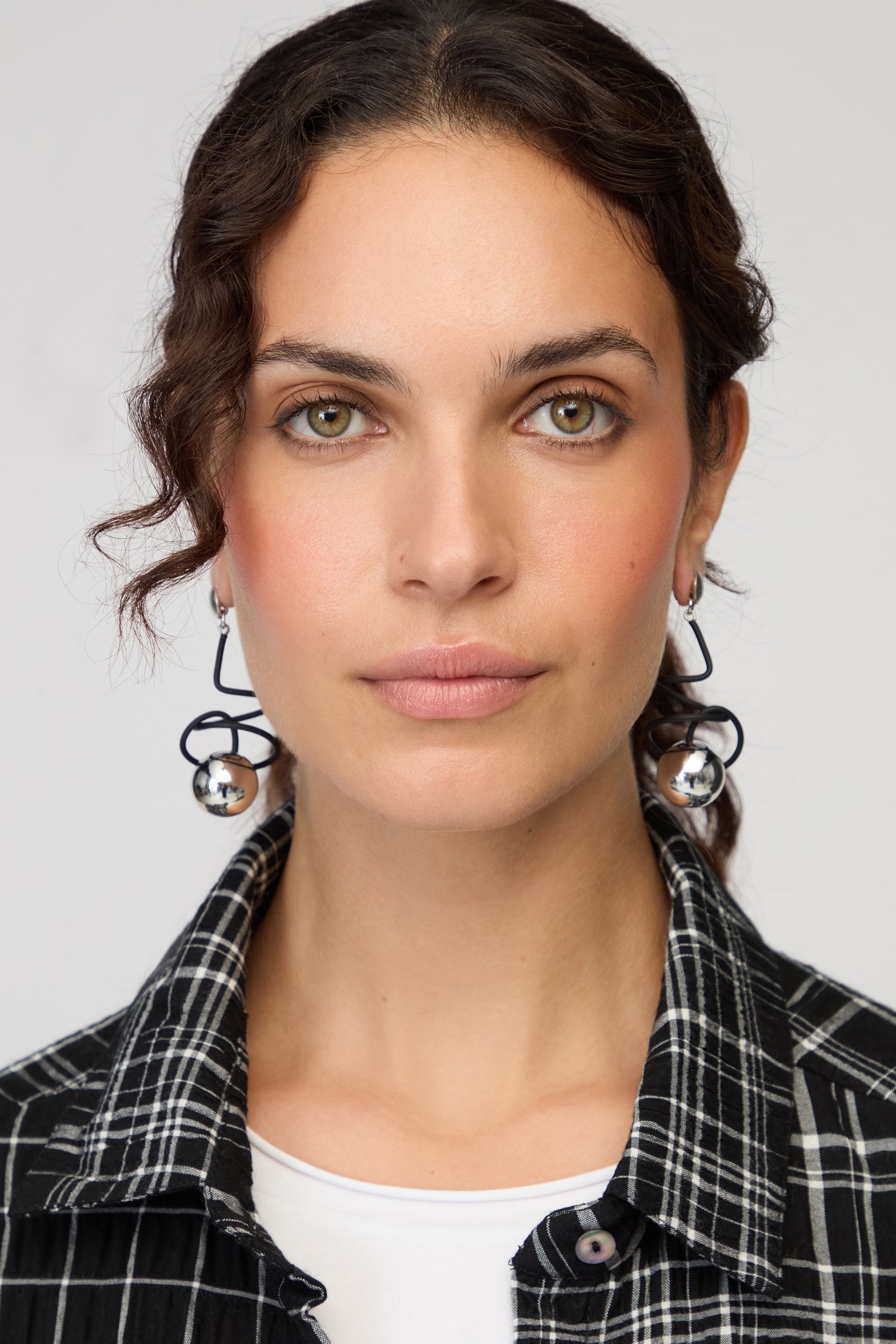 A woman with wavy brown hair, dressed in a black-and-white plaid shirt and white top, wears Graphic Squiggle Earrings and looks directly at the camera against a plain background.