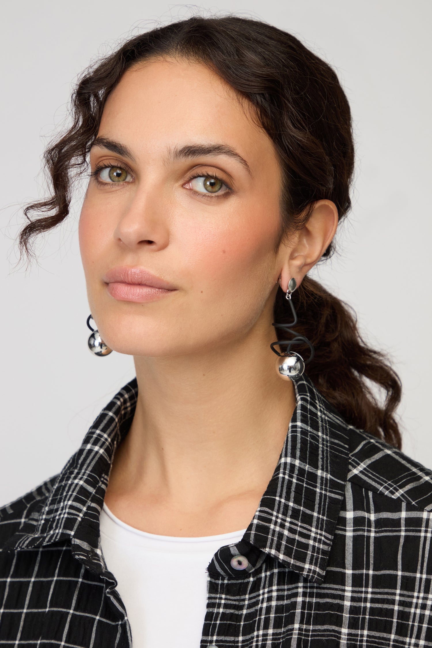 Woman with wavy brown hair wears a black and white plaid shirt, white top, and Graphic Squiggle Earrings inspired by Samuel Coraux, looking at the camera with a neutral expression.