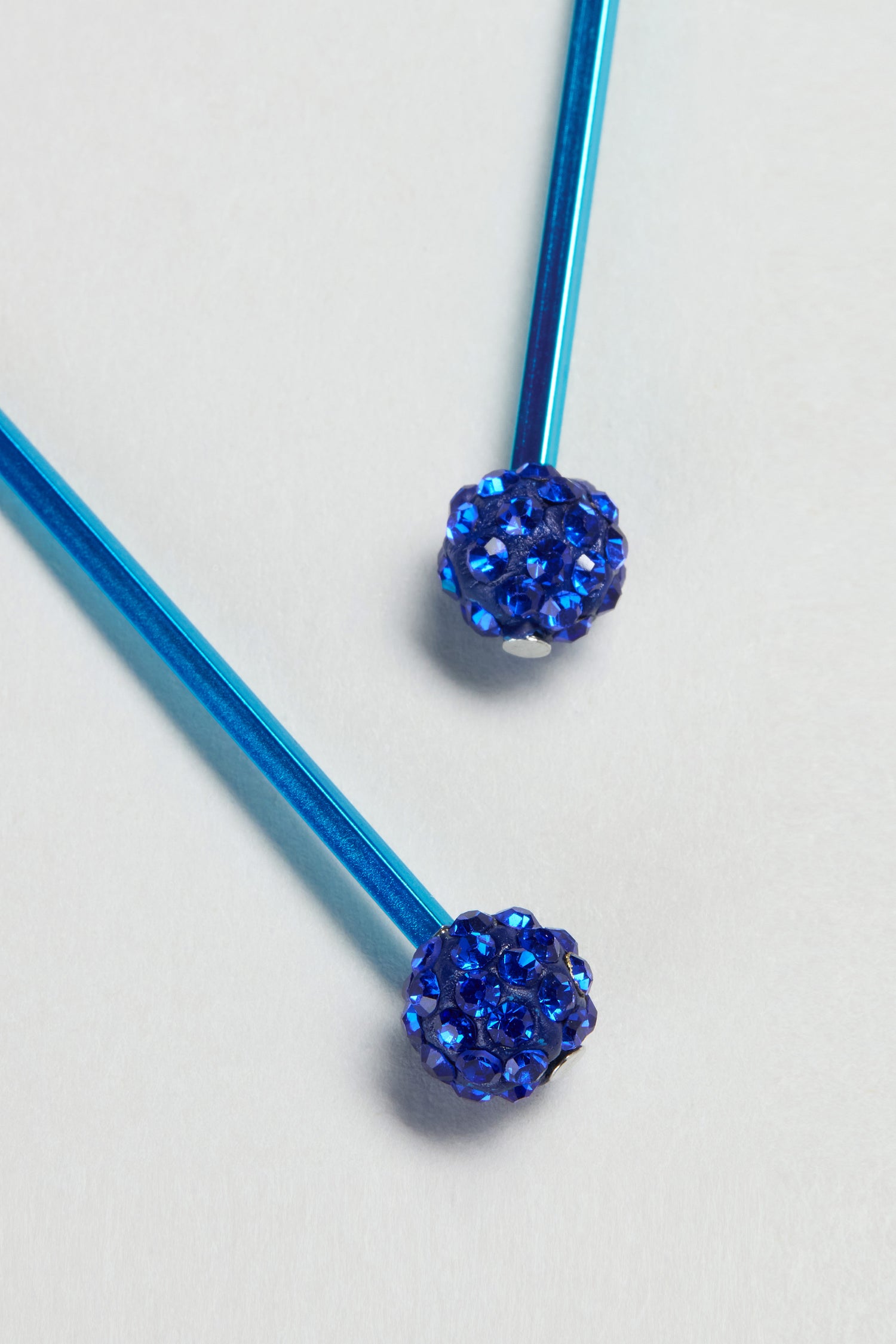 Two blue metallic sticks with round, gem-studded tips are placed on a light surface, reflecting the vibrant flair of Samuel Coraux in these Disco Ball Earrings.