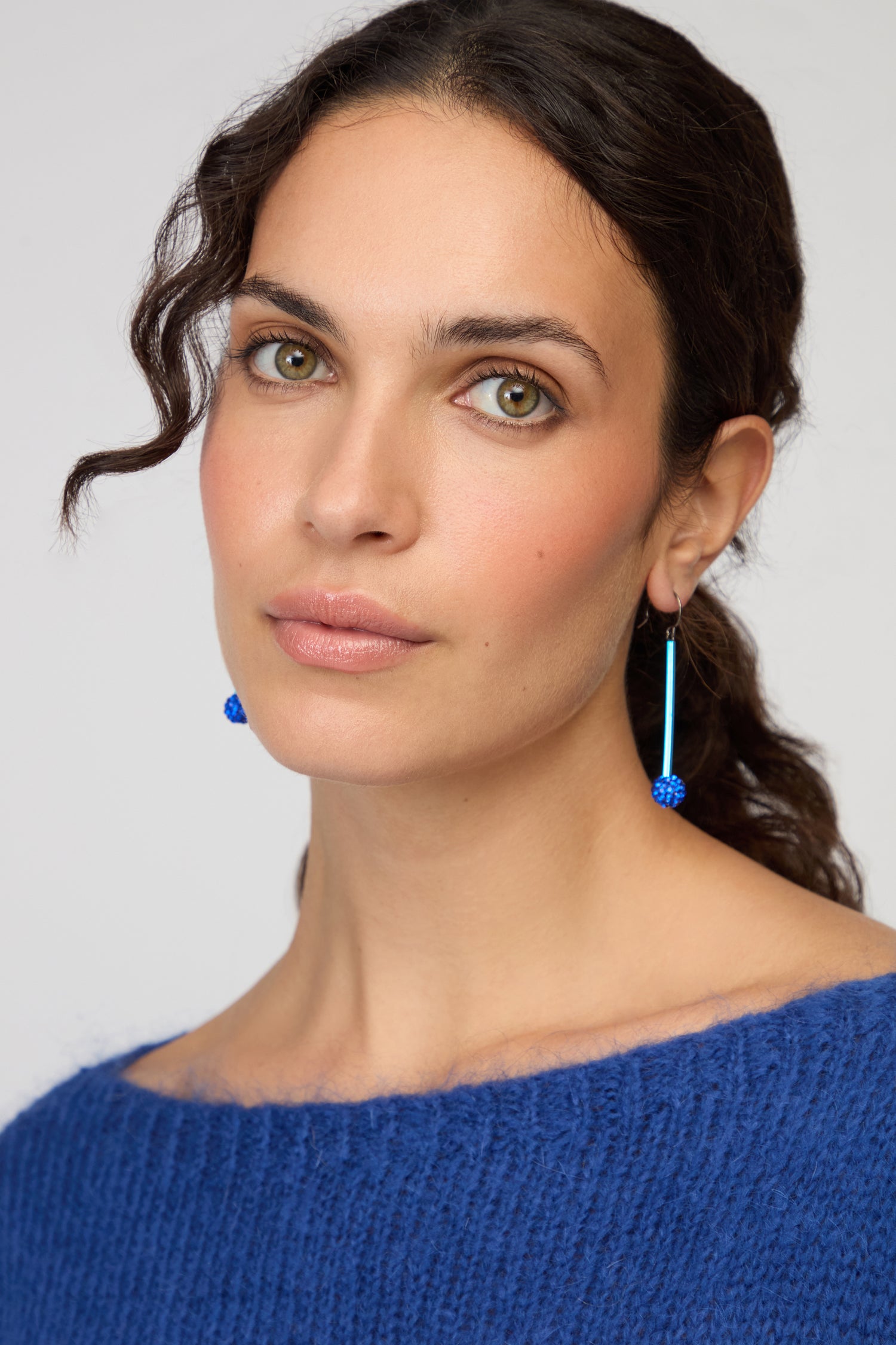 A woman with wavy brown hair wears a blue sweater and striking Disco Ball Earrings by Samuel Coraux. She looks at the camera with a neutral expression against a plain background.
