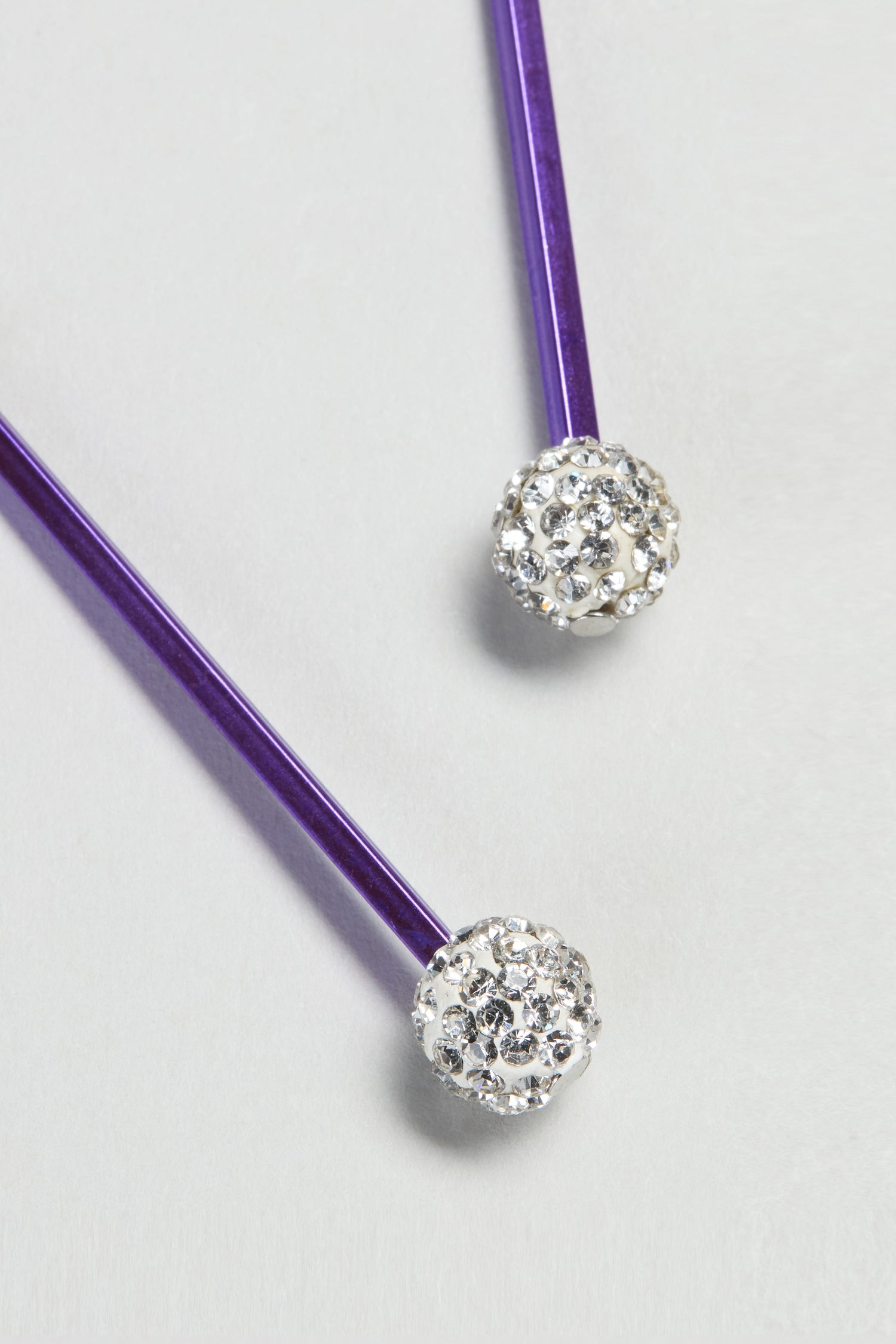 The Disco Ball Earrings feature two purple sticks with round silver ends adorned with clear crystals, capturing a contemporary, playful glamour.