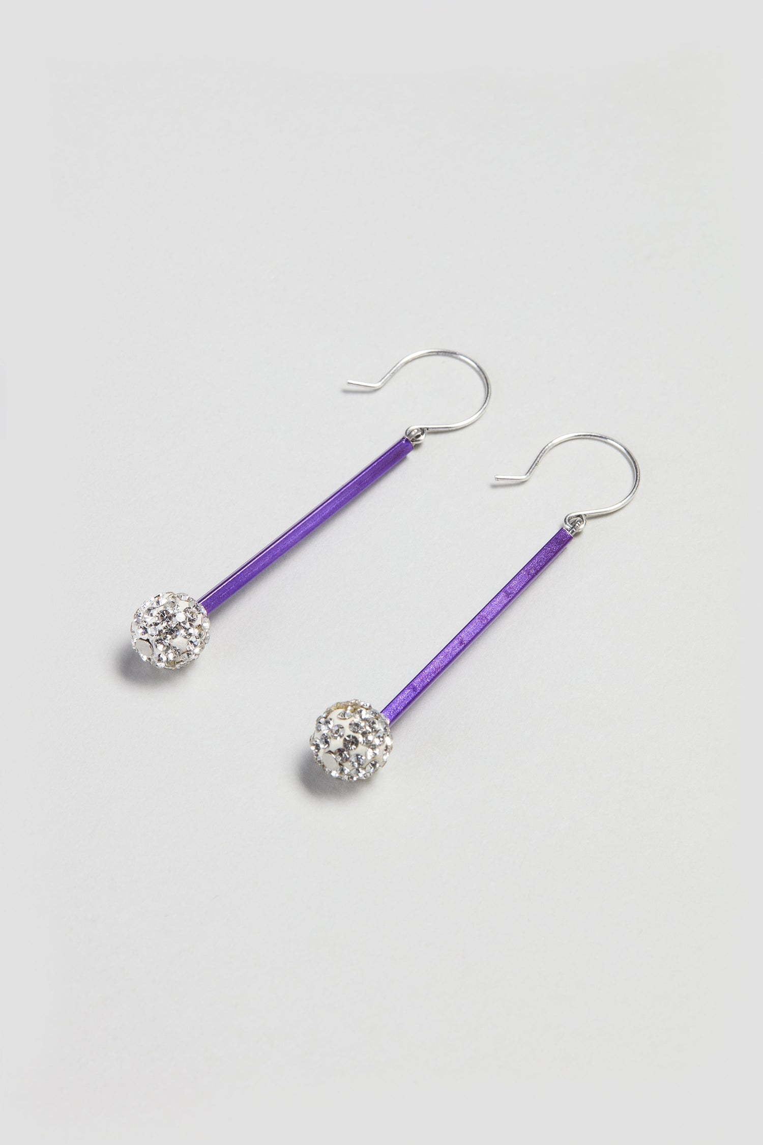 The Disco Ball Earrings feature purple bars and round, faceted clear crystals on silver hooks, offering a stylish contemporary touch. Elegantly displayed on a light background, these earrings enhance any modern jewellery collection.