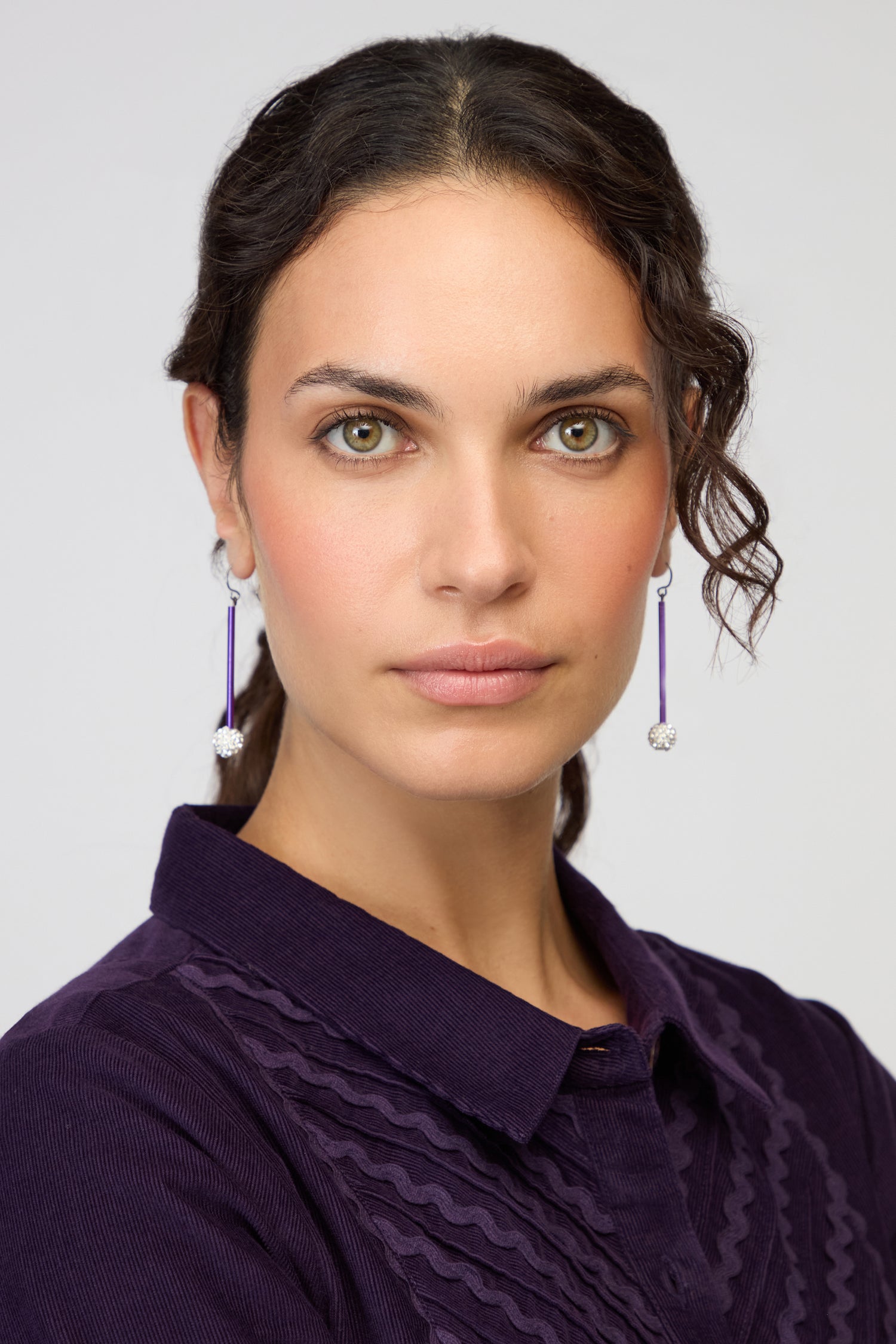 Woman with light eyes and wavy dark hair wears a purple collared shirt and Disco Ball Earrings, facing forward against a plain background—a striking look enhanced by contemporary Disco Ball Earrings.