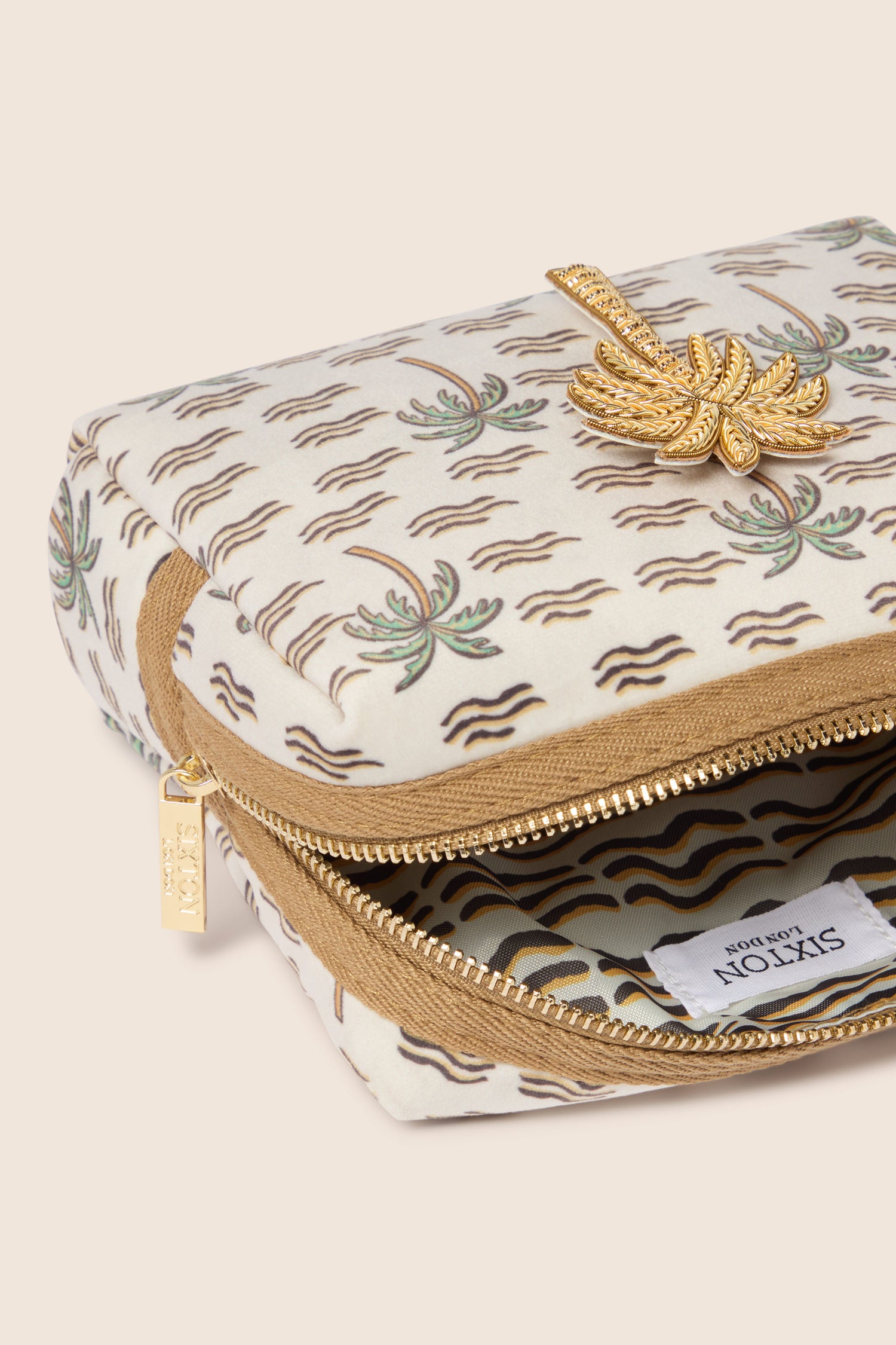 The Large Palm Make-up Bag exudes tropical elegance with its white exterior adorned with brown and green palm tree patterns. The bag features a zipper accented by a decorative gold palm tree design. Partially open, it reveals a white interior decorated with wavy lines.