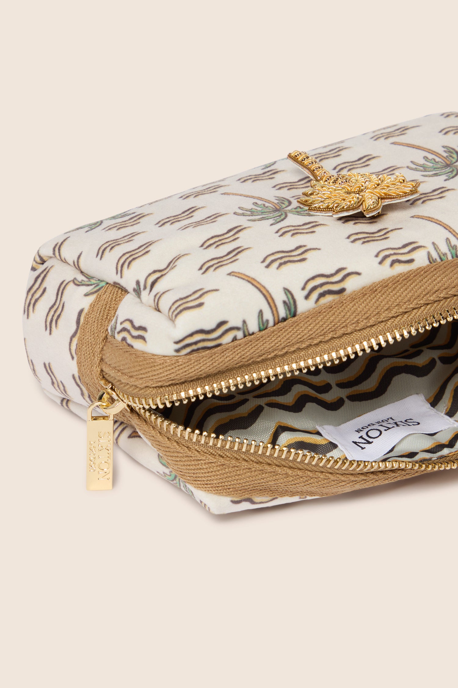 A close-up of the Small Palm Make-up Bag with a partially open zipper, revealing a lined interior. This beige and patterned bag, reminiscent of tropical vibes, features intricate gold detailing and an attached gold tag—ideal for holding your beauty essentials.