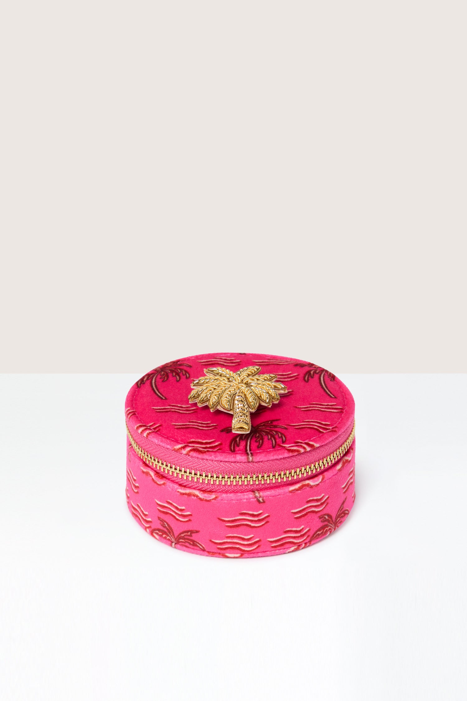 The Palm Jewellery Travel Pot is a round pink fabric box adorned with a golden embroidered pattern and equipped with a gold zipper. A gold bee decoration graces the lid, and inside, a chic zebra print lining adds an unexpected flair, making it the ideal travel container for your jewellery.