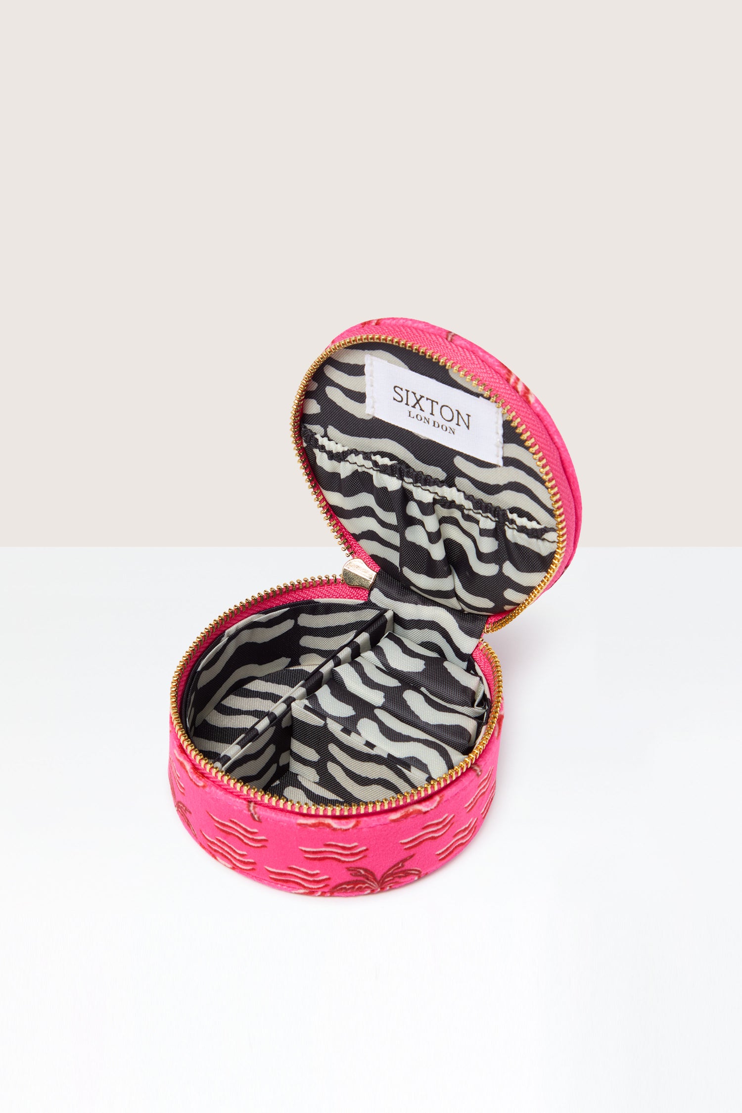 The Palm Jewellery Travel Pot is a round, pink travel accessory featuring a gold zipper, golden accents, and a chic zebra print lining. Inside, the label reads "Sixton London.