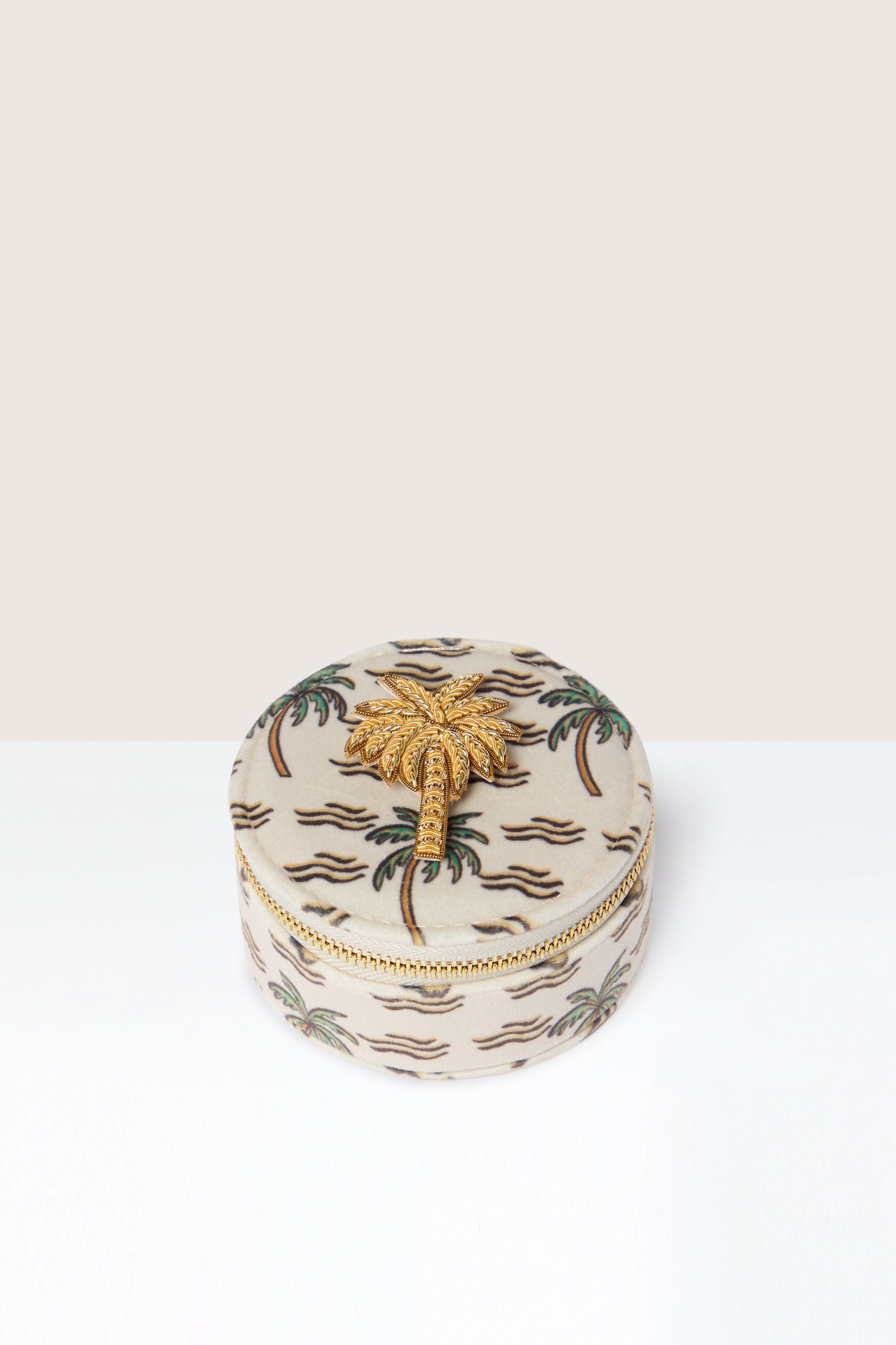 The Palm Jewellery Travel Pot is a round, fabric-covered jewelry case adorned with a pattern of palm trees and featuring a zebra print lining. The lid boasts a gold-tone, palm tree-shaped clasp that adds a touch of elegance to the gold palm tree design.