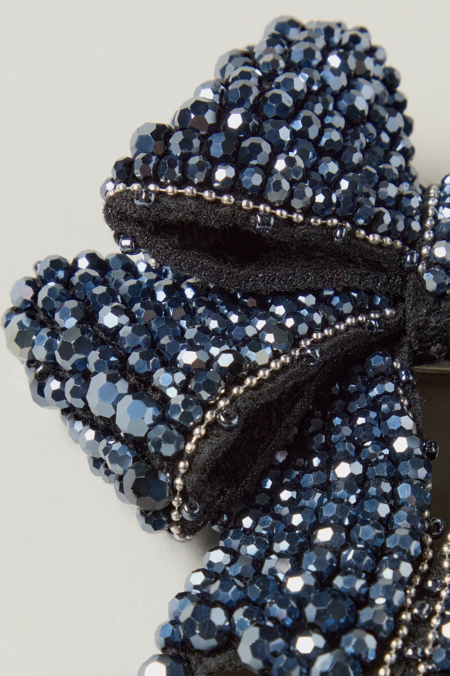 Close-up of the Hand Beaded Bow Brooch, adorned with shiny dark blue sequins and beads on a black fabric base, set against a light background.