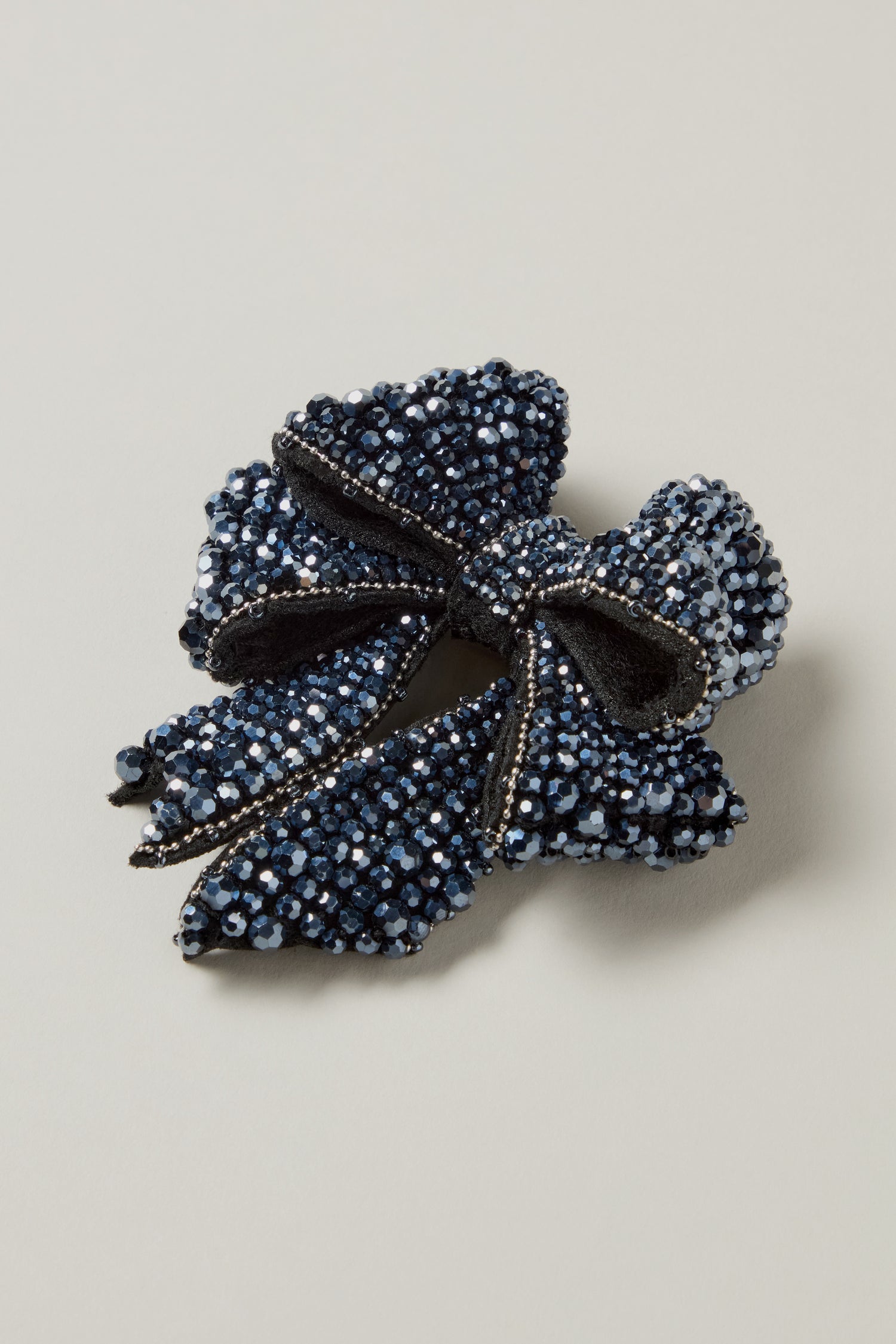 The Hand Beaded Bow Brooch features navy blue velvet, hand-embellished with sparkling beads and rhinestones, shown on a simple light background.