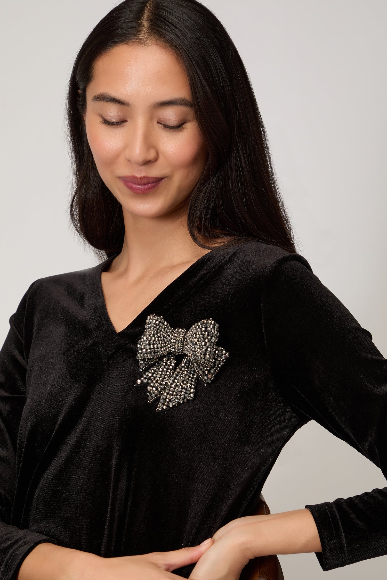 A woman with long dark hair wears a black velvet top, accessorized with the Hand Beaded Bow Brooch, and looks down with a slight smile.