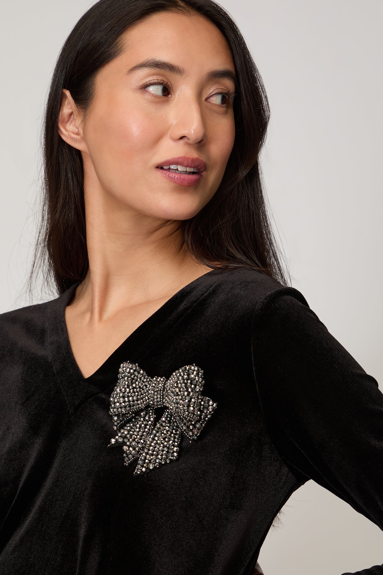 A woman looks to the side against a neutral background, wearing a black velvet top adorned with the Hand Beaded Bow Brooch on her chest.