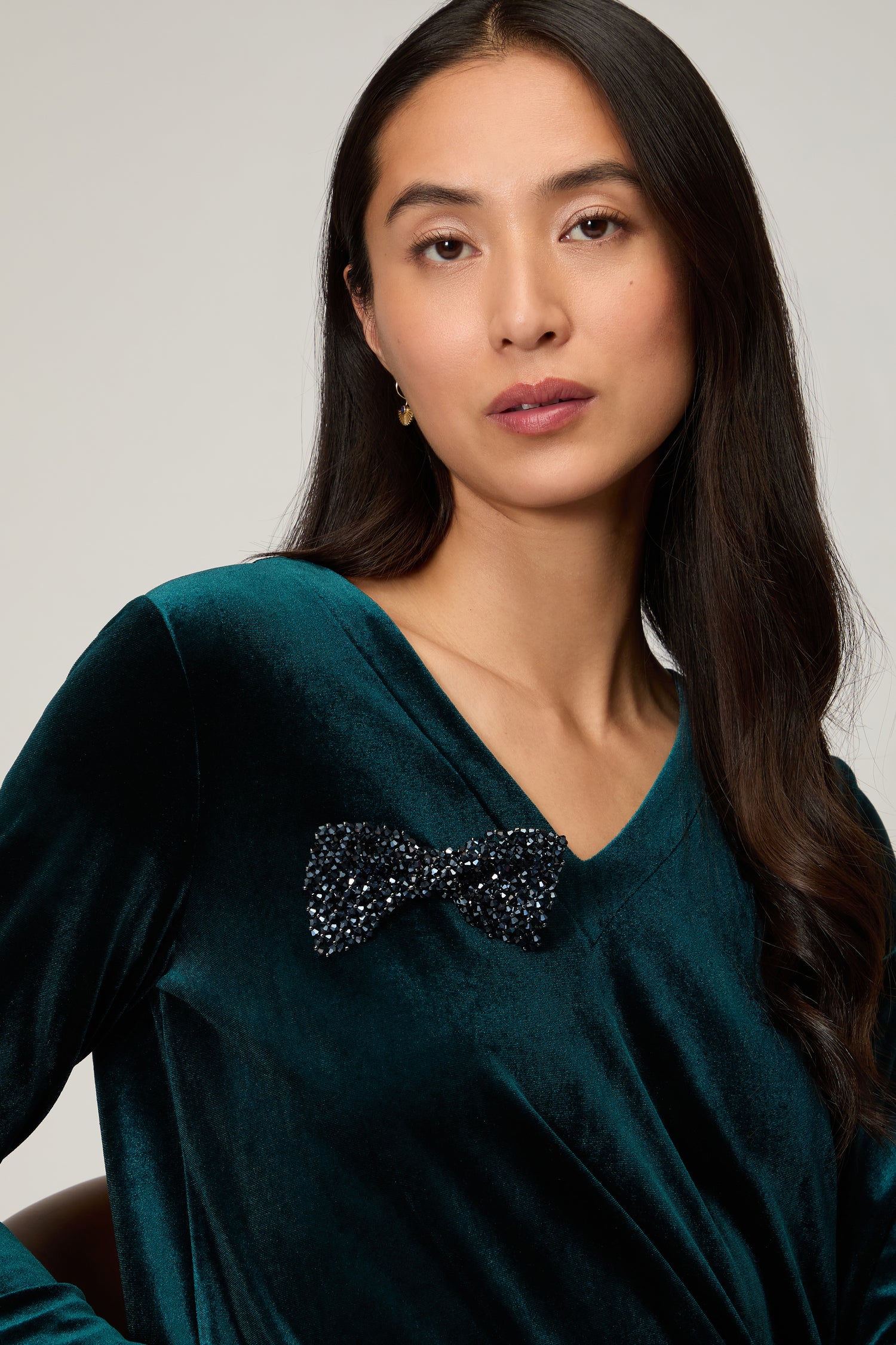 A woman with long dark hair wears a dark green velvet top styled with the Hand Beaded Bow Tie Brooch—a black, beaded bow accessory on her chest that adds a modern, versatile accent to her look.