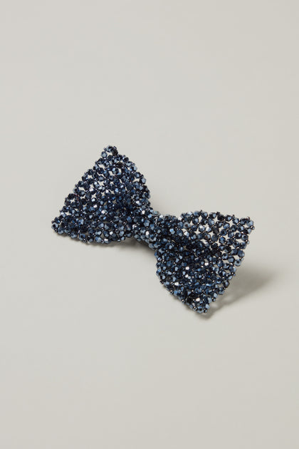 The Hand Beaded Bow Tie Brooch is navy blue with small, reflective crystals and is shown on a plain light grey background.