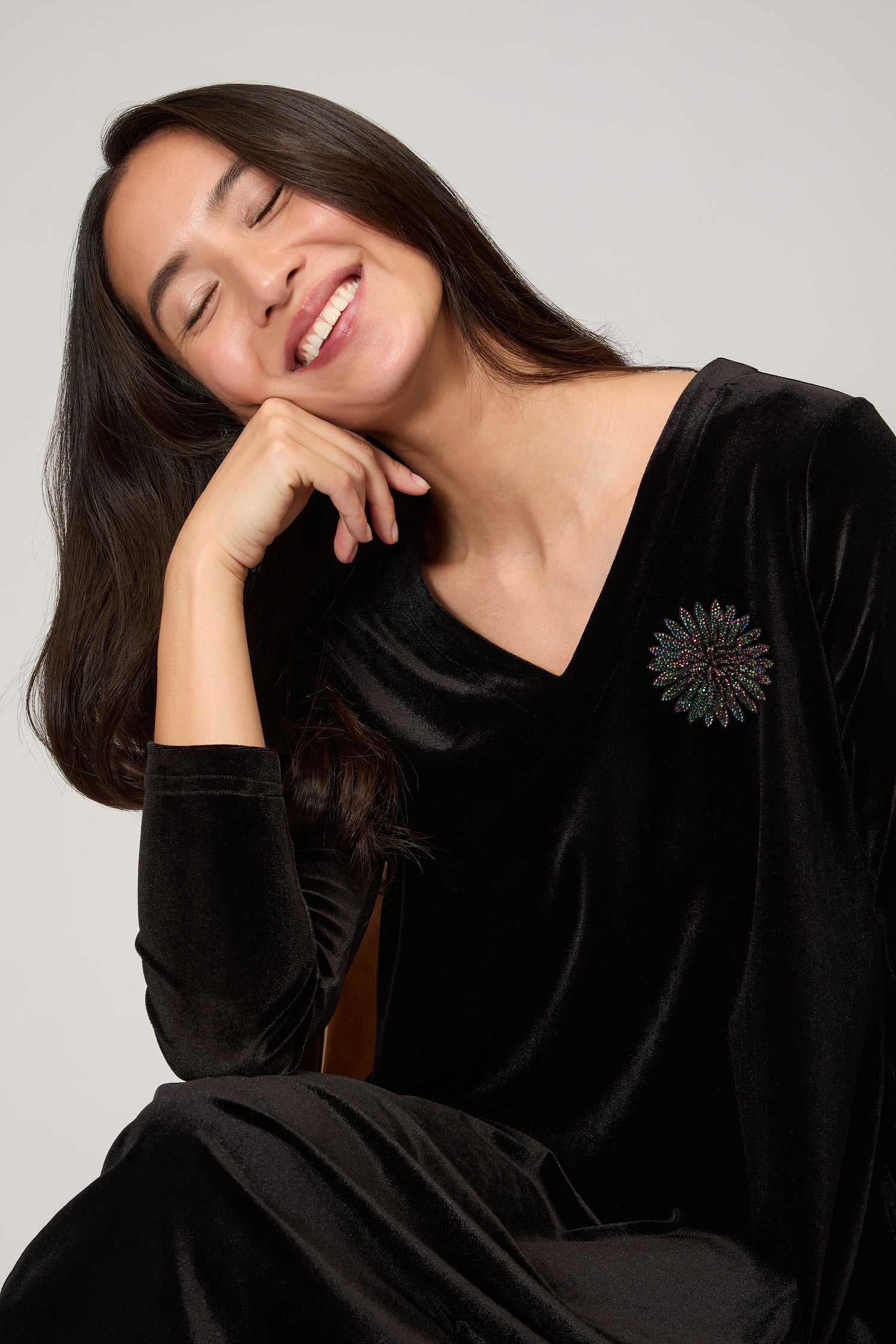 A woman with long dark hair smiles with closed eyes, resting her chin on her hand, wearing a black velvet dress accented by the Dahlia Flower Brooch on her chest.