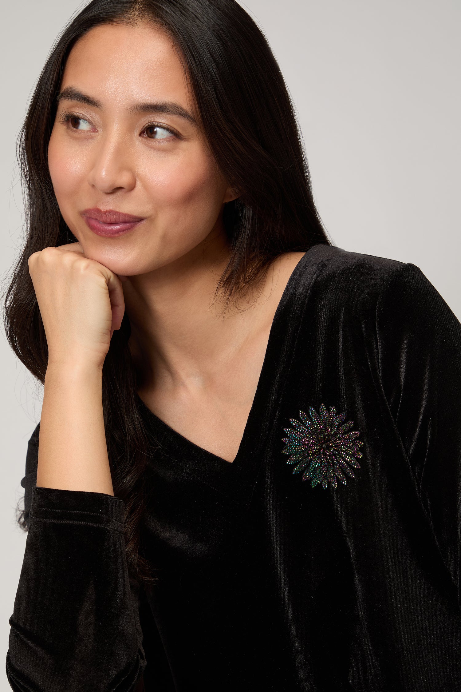 Woman in a black V-neck velvet top wears the Dahlia Flower Brooch, her floral accessory, as she looks to the side with her chin resting on her hand.