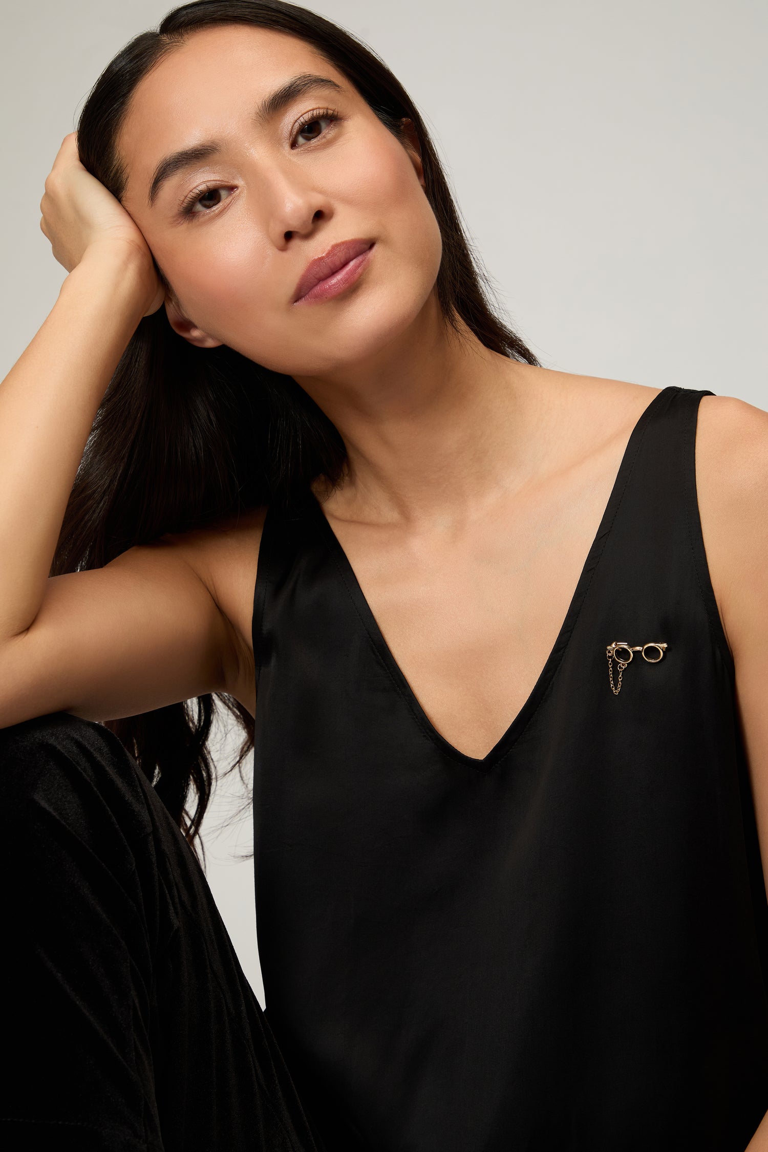 A woman with long dark hair rests her head on her hand, wearing a sleeveless black top adorned with the playful Spectacles Brooch on the left side.