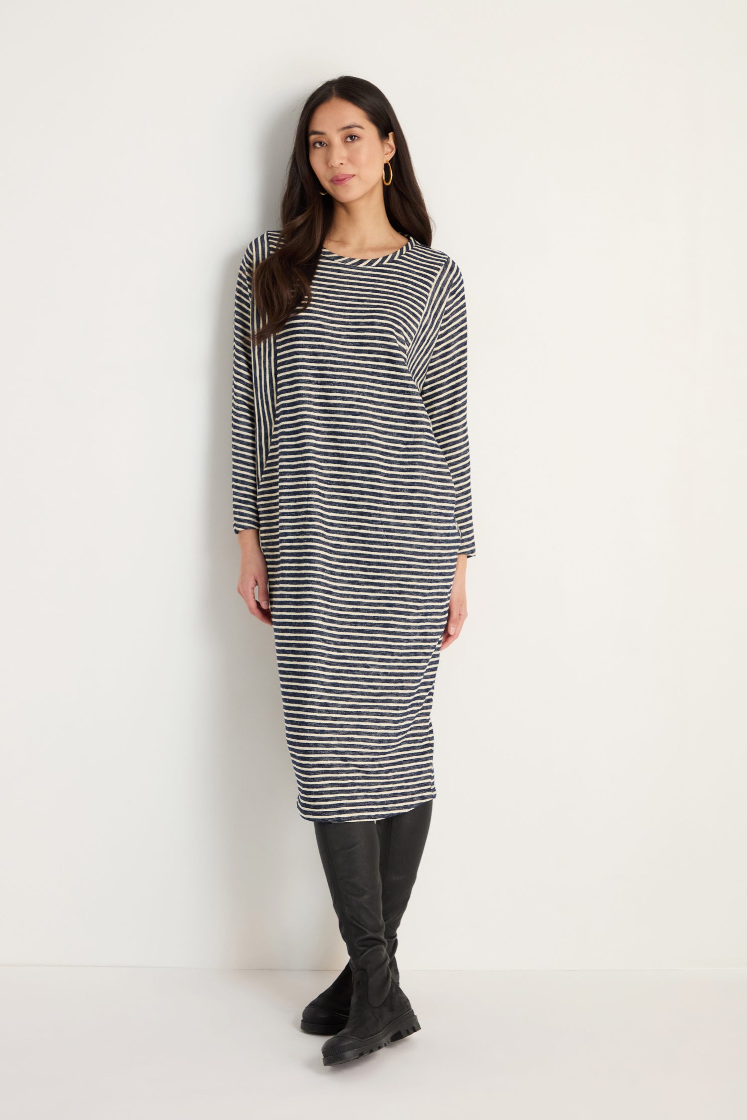A woman stands against a white wall wearing the Textured Stripe Jersey Dress, which features black and white stripes, long sleeves, and is paired with black knee-high boots.