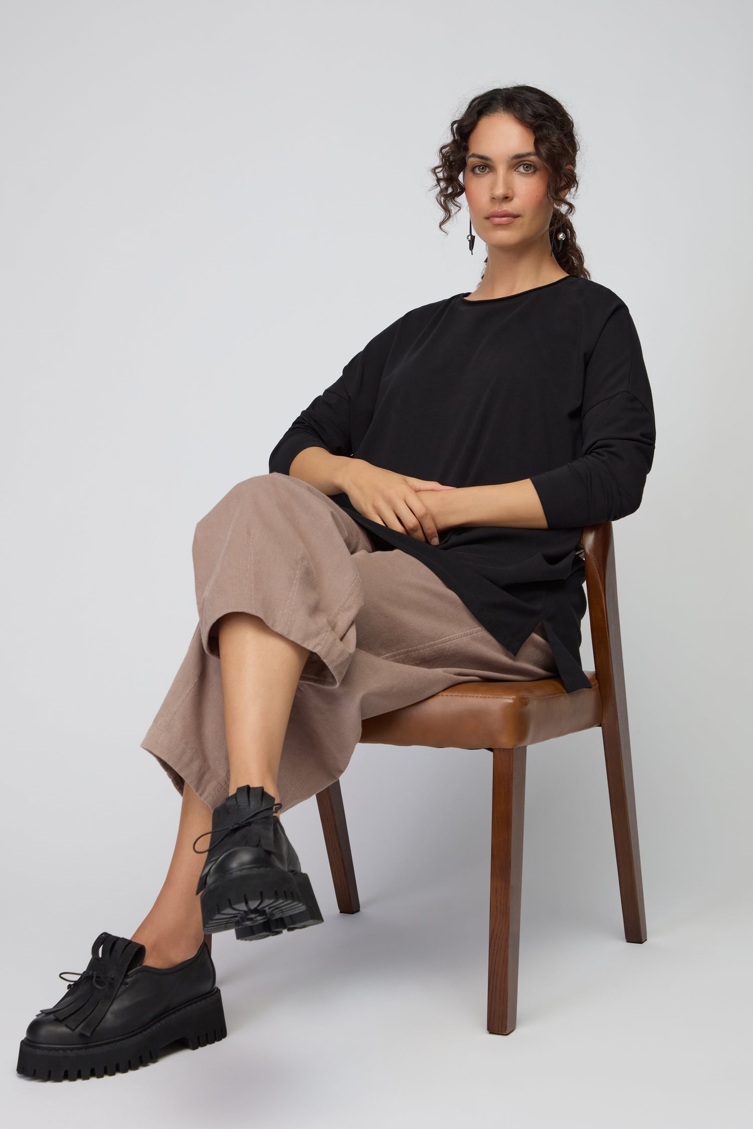 A woman with curly hair sits on a wooden chair, wearing the Cotton Jersey Relaxed Top in black, paired with beige wide-leg pants and black platform shoes against a plain background.