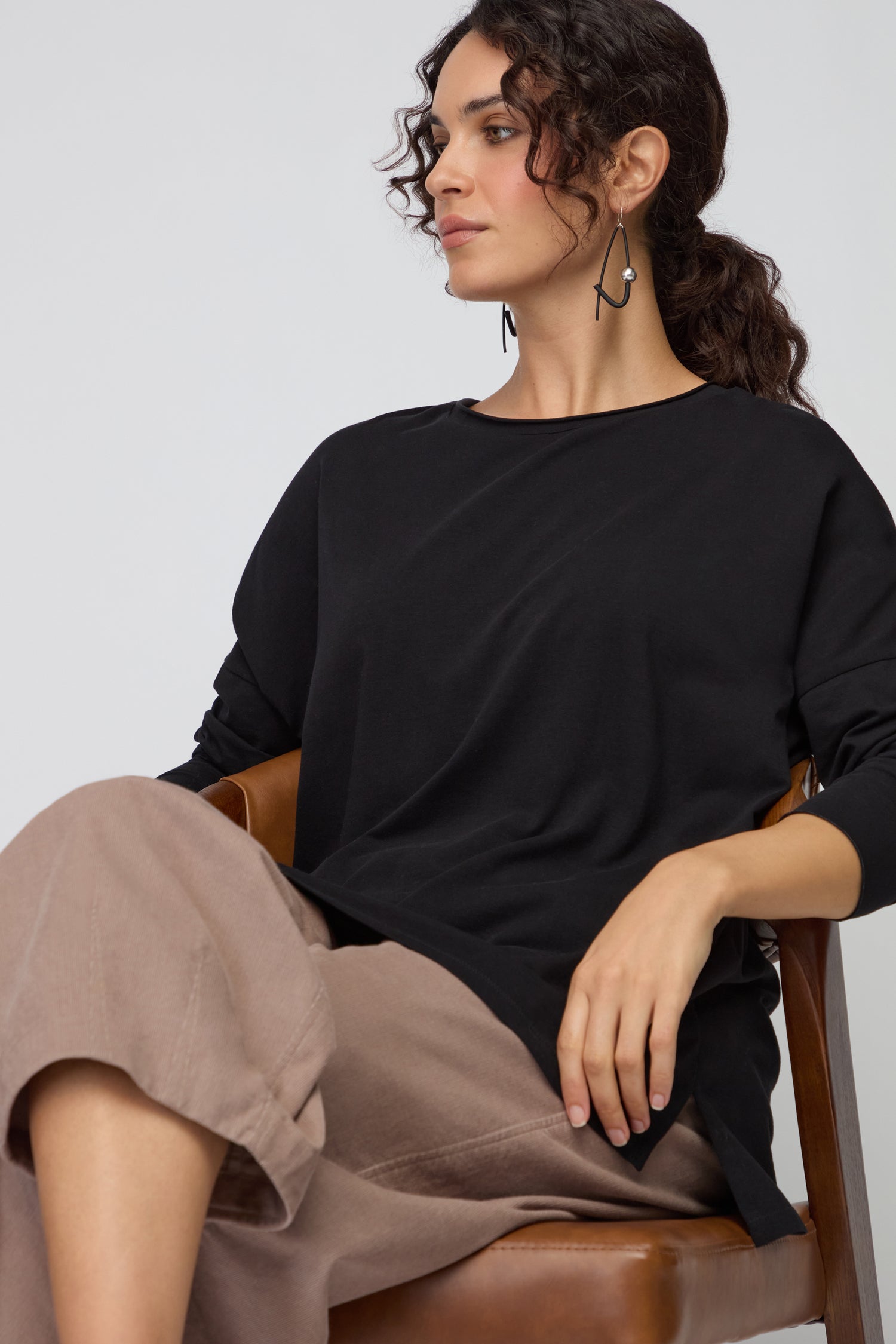 A woman with curly hair sits on a brown chair, wearing the Cotton Jersey Relaxed Top in black—an effortless wardrobe essential—paired with beige pants and dangling earrings, looking to the side.