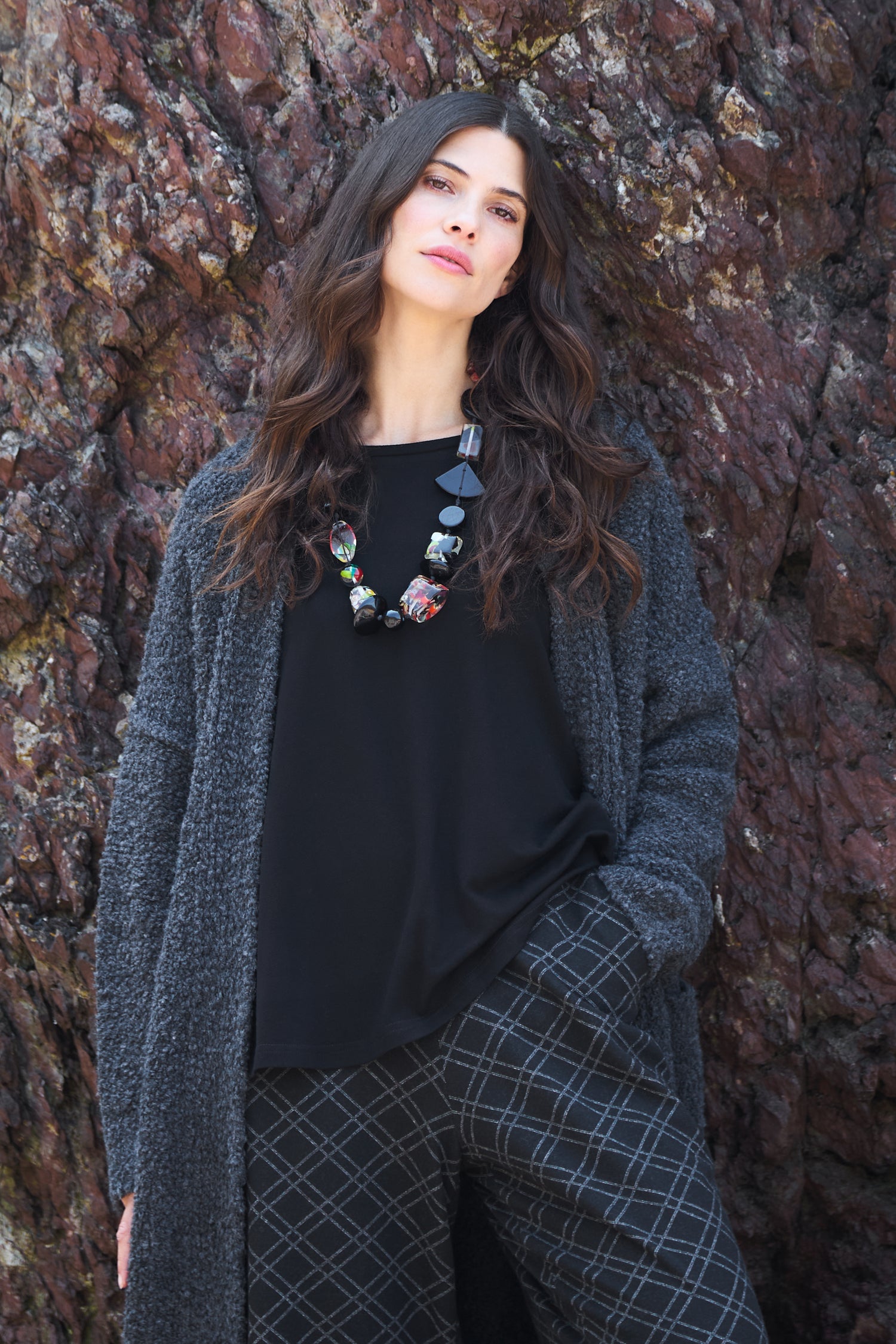 A woman with long dark hair stands against a rocky background, wearing a grey textured coat, plaid pants, a chunky beaded necklace, and the Cotton Jersey Relaxed Top—a true wardrobe essential.