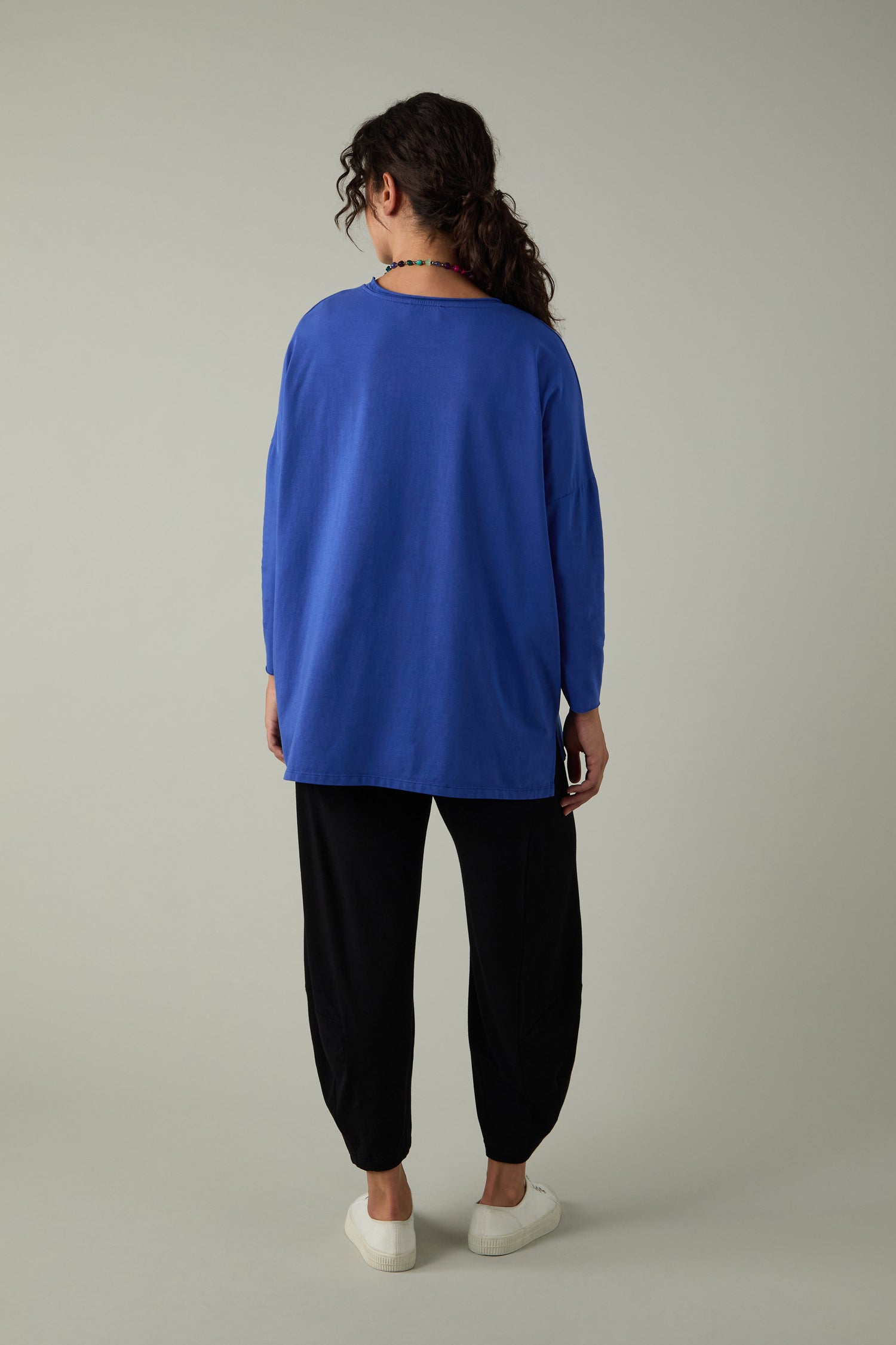 Person stands facing away, dressed in a blue Cotton Jersey Relaxed Top with a relaxed fit, paired with black pants and white shoes against a plain light gray backdrop—this top is the ultimate wardrobe essential.