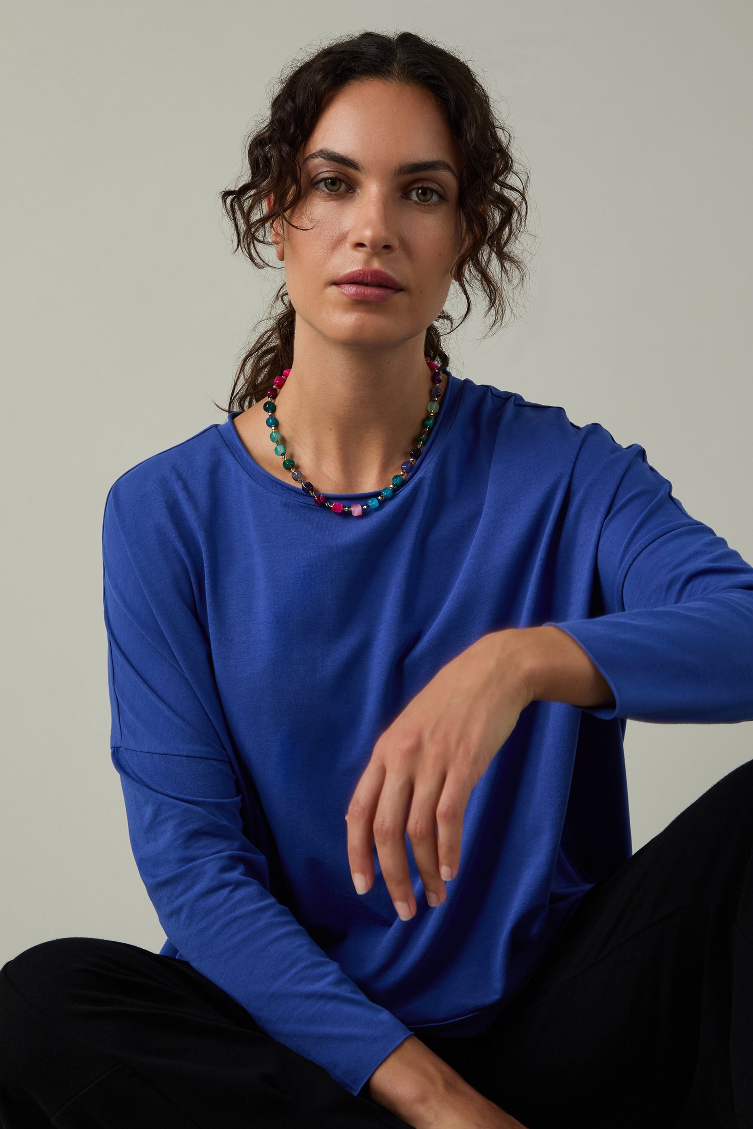 A person with curly hair, dressed in a blue Cotton Jersey Relaxed Top and black pants with a colorful beaded necklace, sits against a neutral background, showcasing effortless style and the relaxed fit of the Cotton Jersey Relaxed Top.