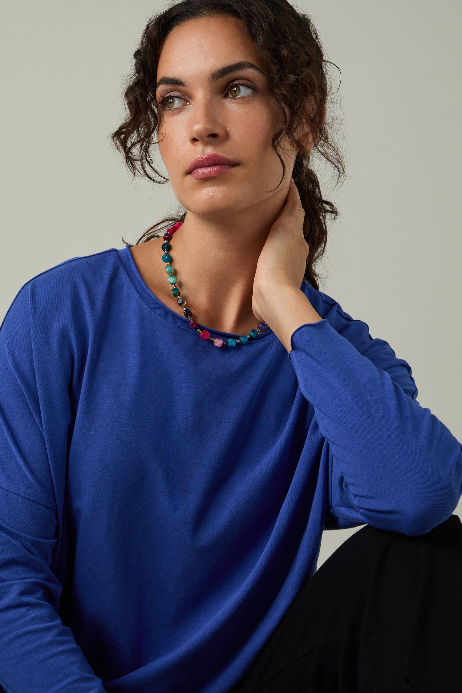 A woman with curly hair wears a blue Cotton Jersey Relaxed Top and a colorful beaded necklace, sitting with her hand on her neck and looking to the side.